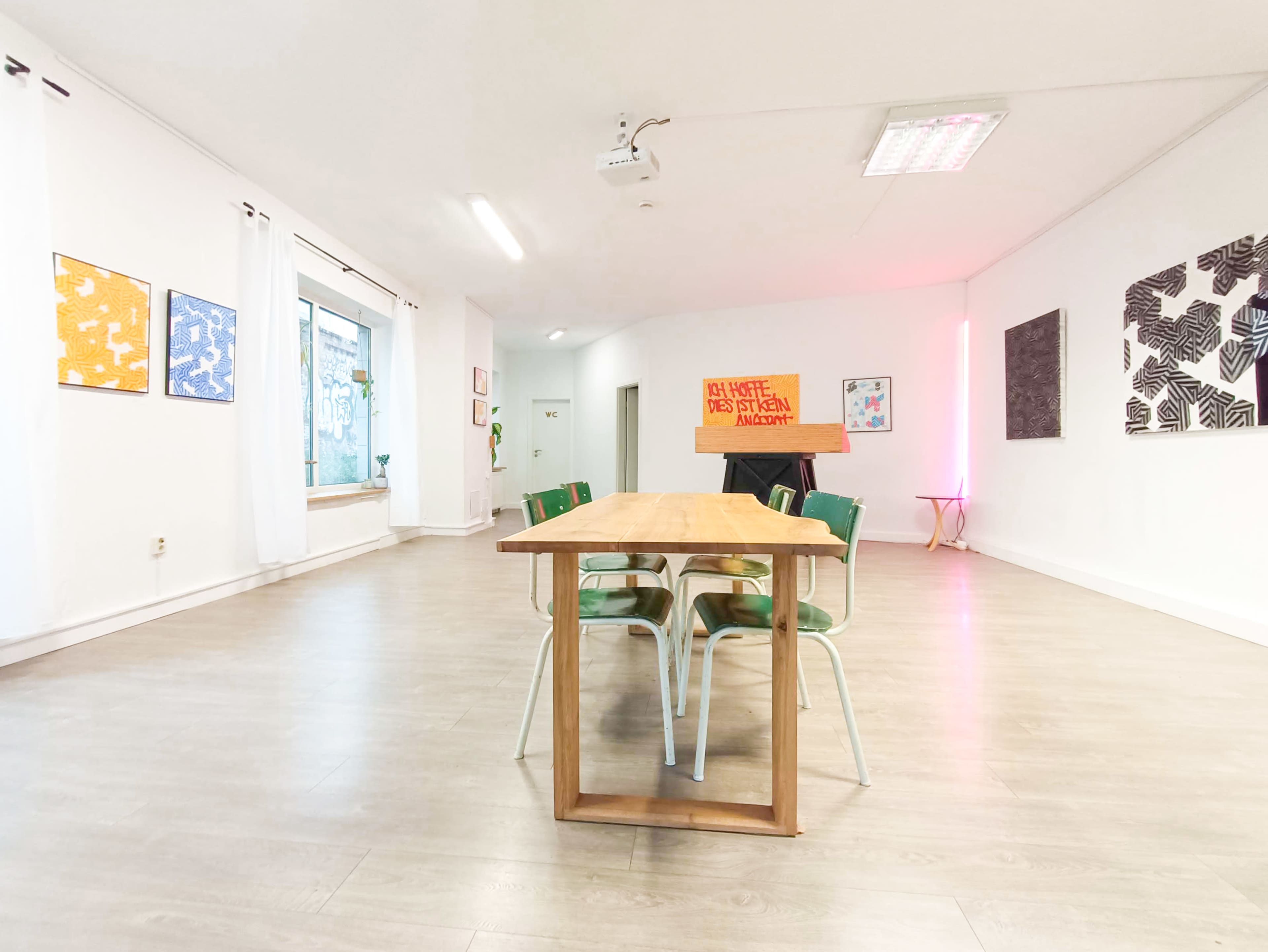 The image shows a spacious room with a wooden table surrounded by green chairs, adorned with colorful artwork on the walls.