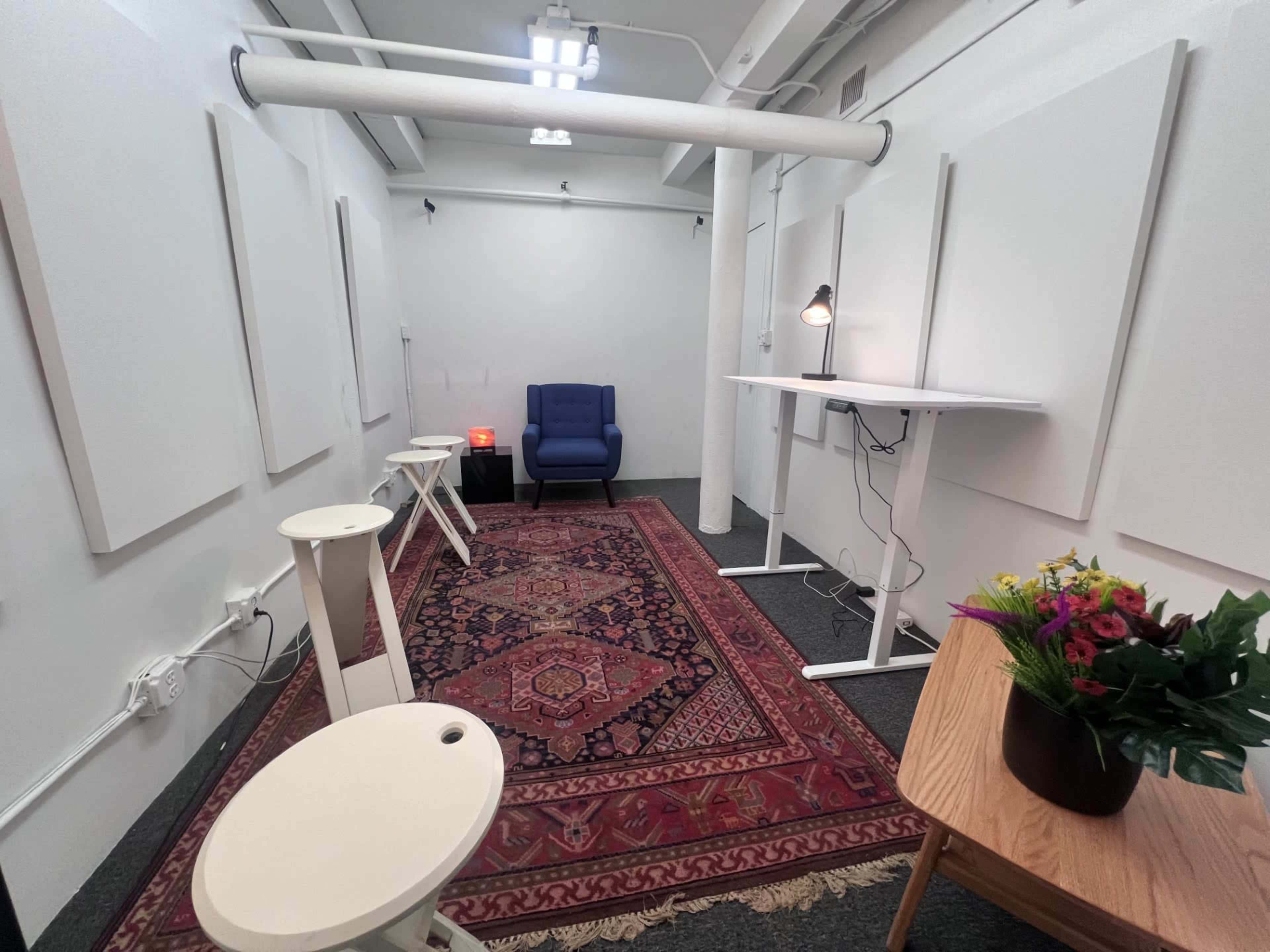 The image shows a small room with white walls, a blue chair, a desk, two small stools, a lamp, and a rug with a floral arrangement on a table.