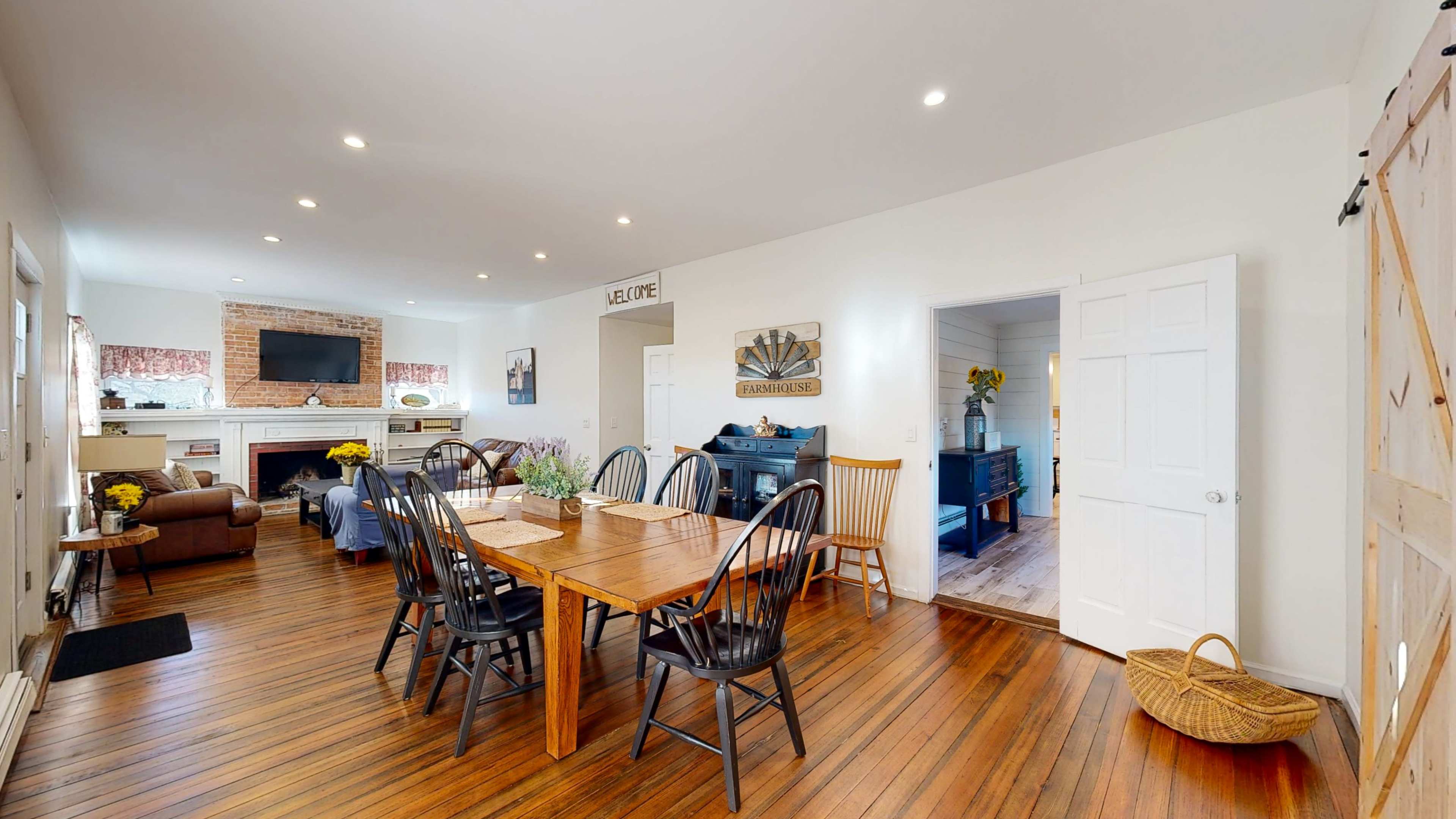 A spacious dining area with a wooden table surrounded by black chairs, leading into a living space with a brick fireplace and a doorway to another room.
