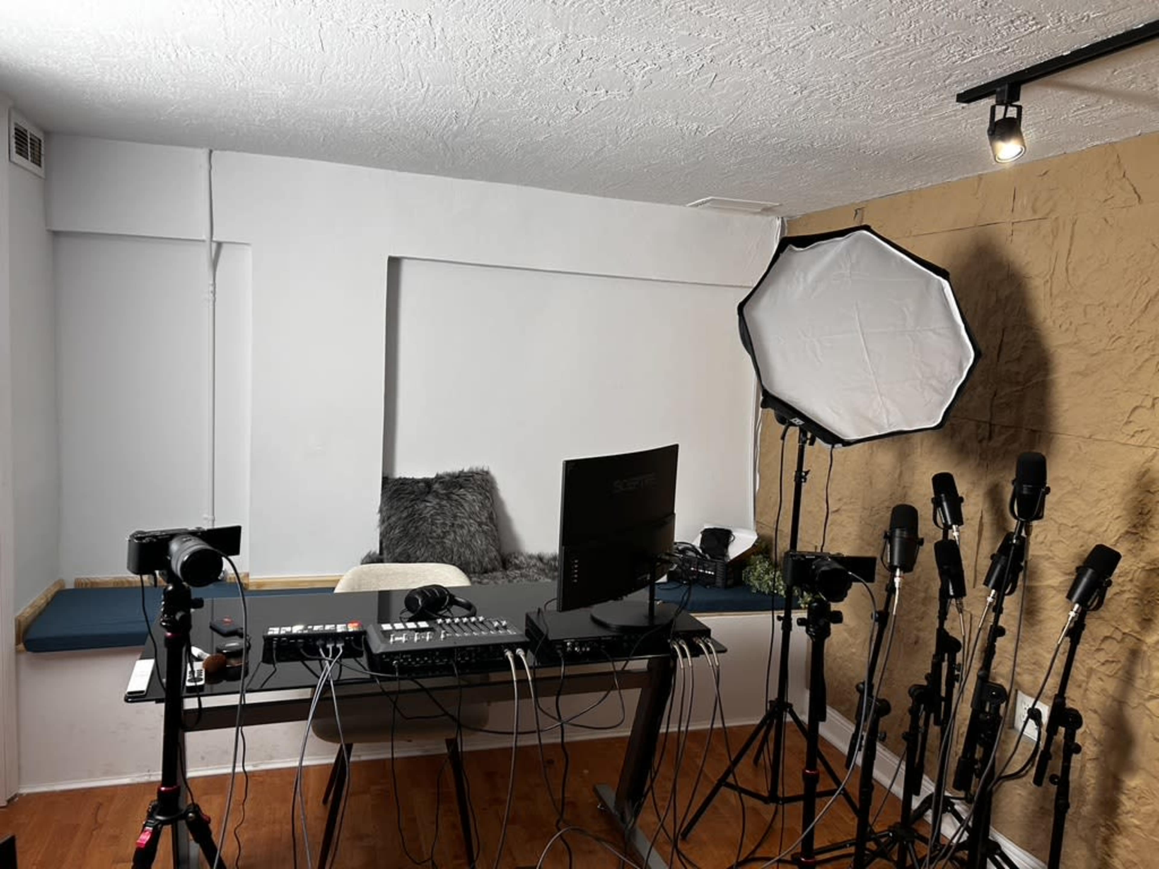 A recording studio setup featuring a large desk with a computer, multiple cameras, microphones on stands, and a softbox light.