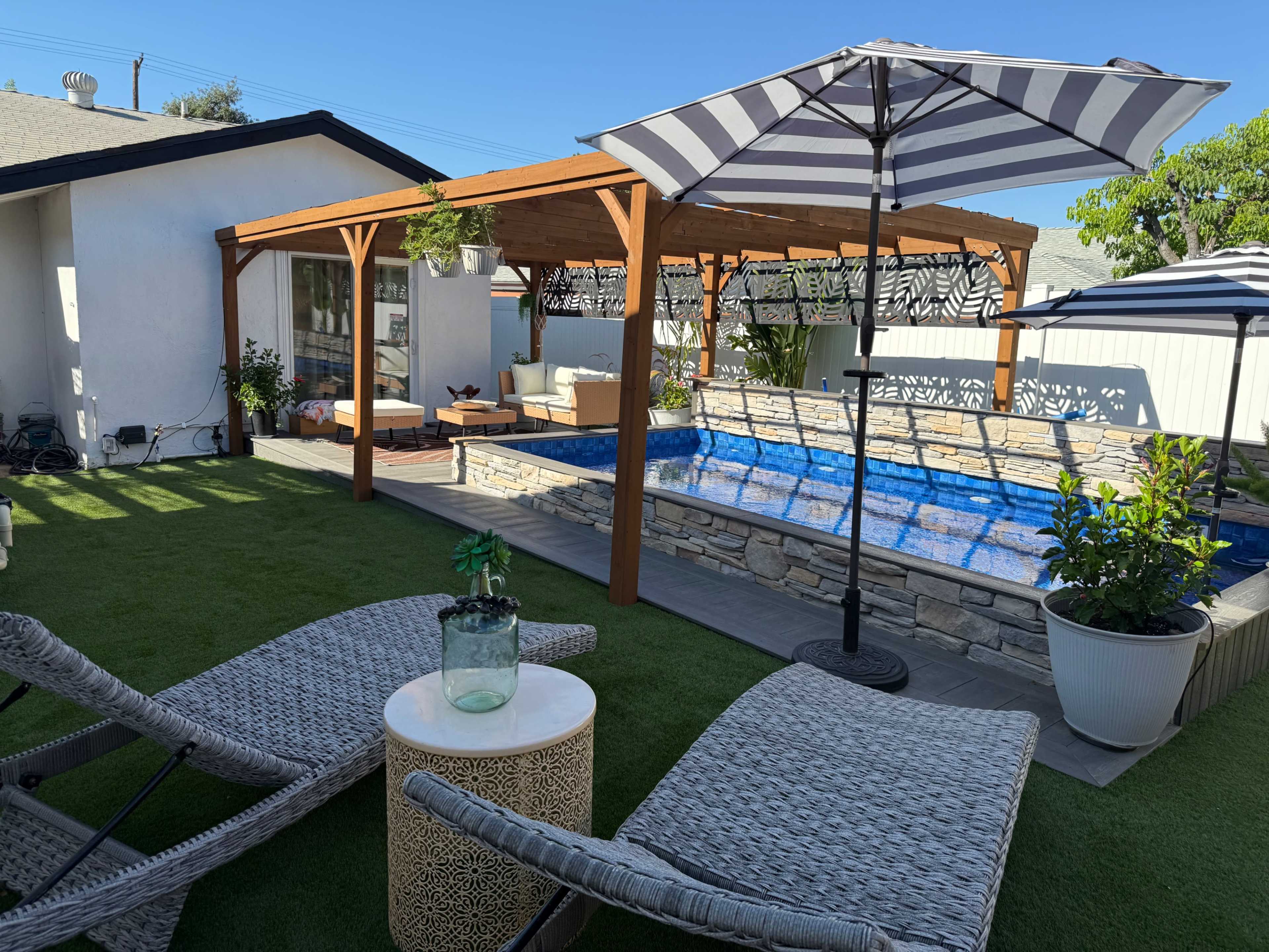 The image shows a backyard with a swimming pool, wooden pergola, and lounge chairs surrounded by artificial grass and potted plants.