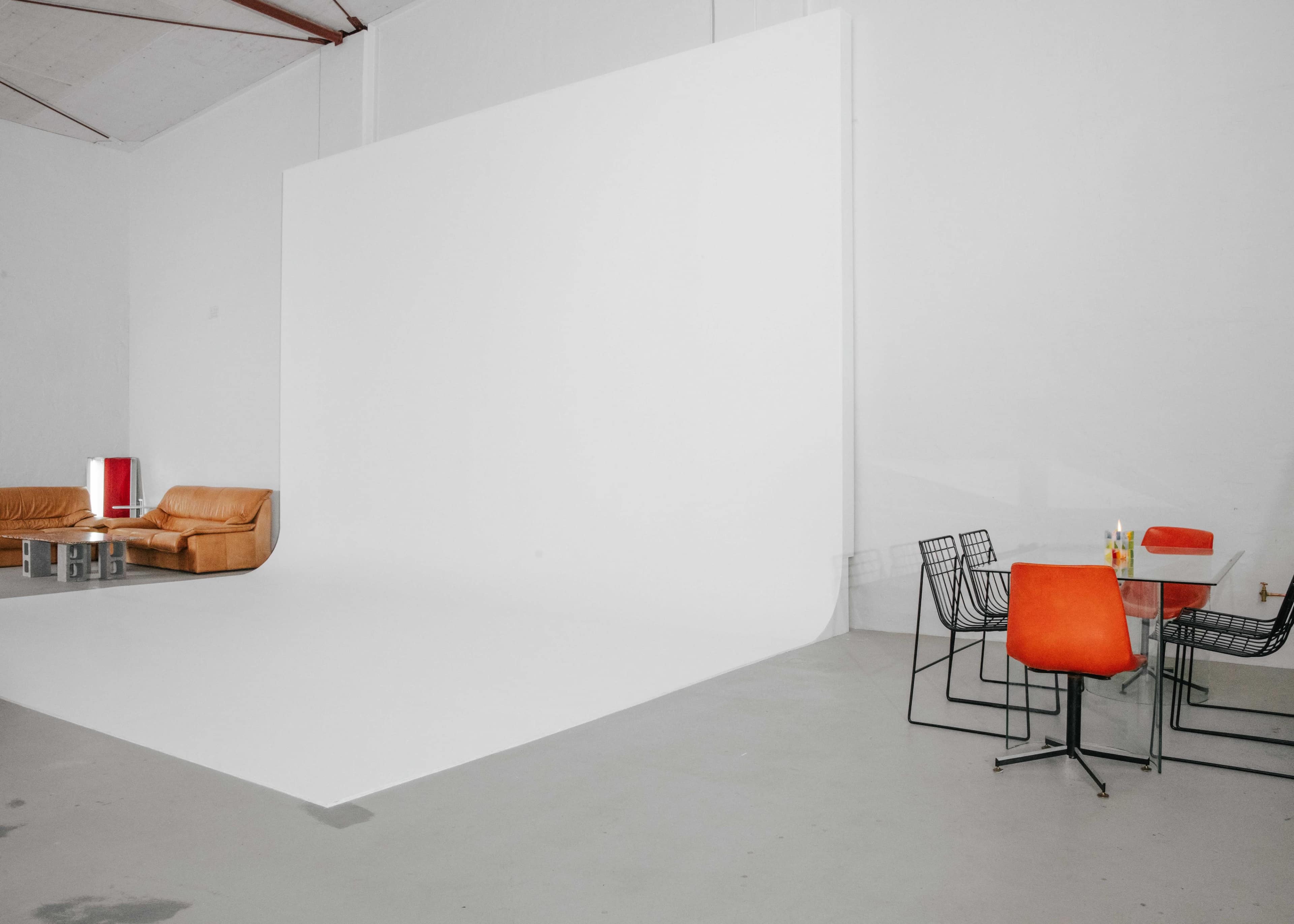 A minimalist studio space featuring a white backdrop, an orange sofa, a glass table with orange chairs, and a few drinks on the table.