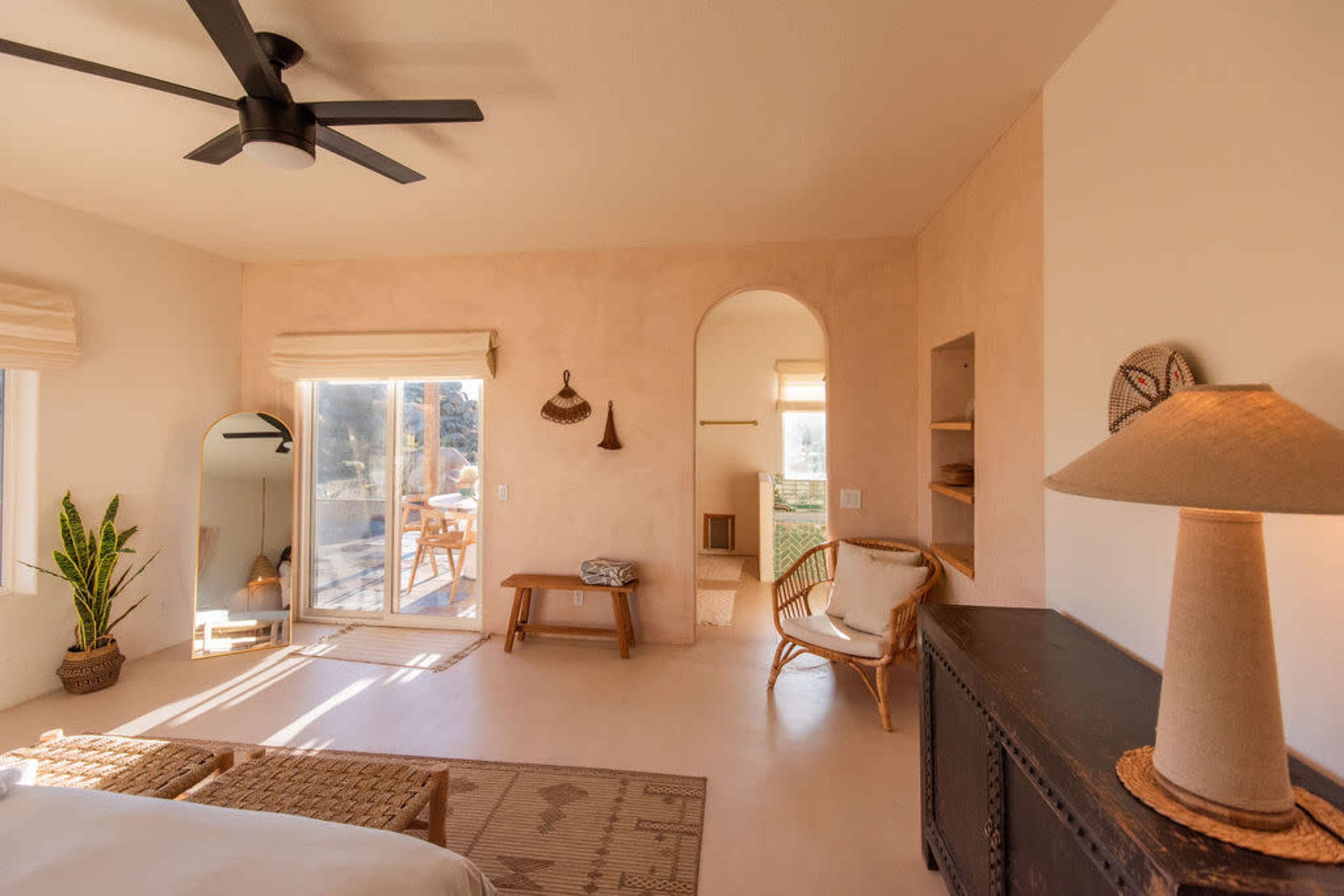 A minimalist bedroom features beige walls, a ceiling fan, natural light from sliding glass doors, and simple furnishings, including a chair and a table.