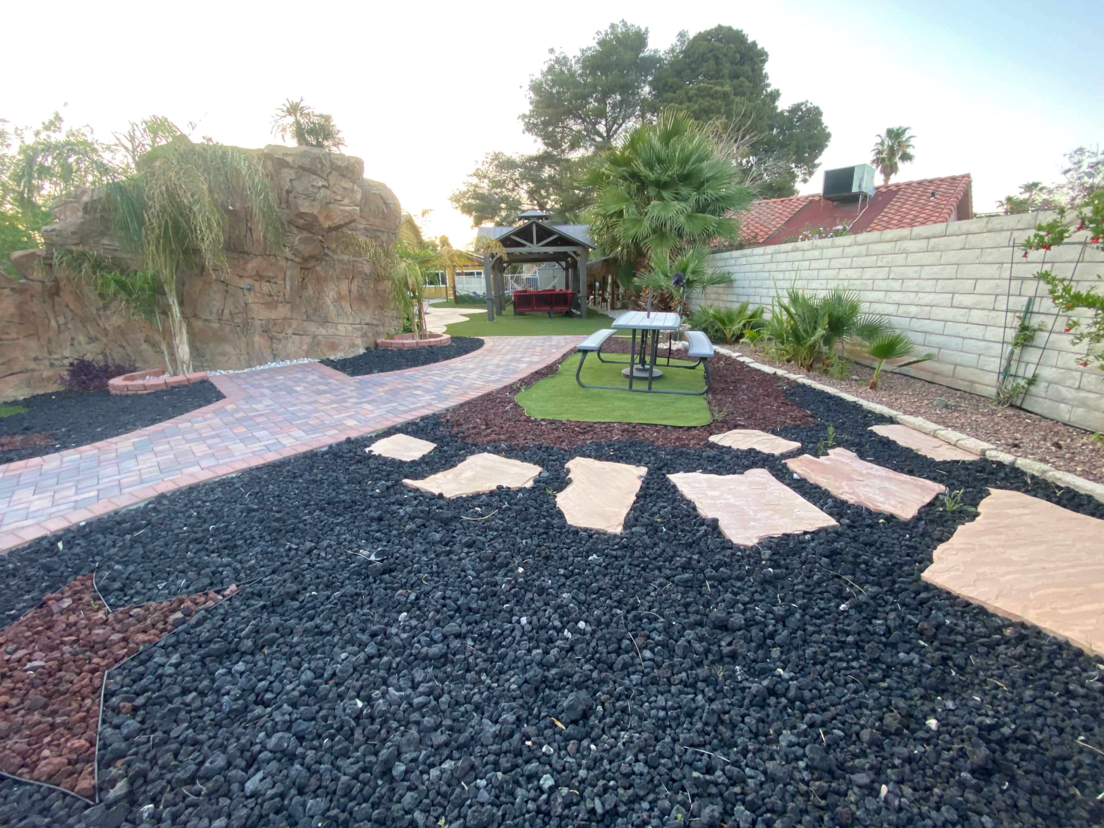 The image shows a landscaped backyard featuring a stone pathway, artificial grass, and decorative rocks, with a pavilion in the background.