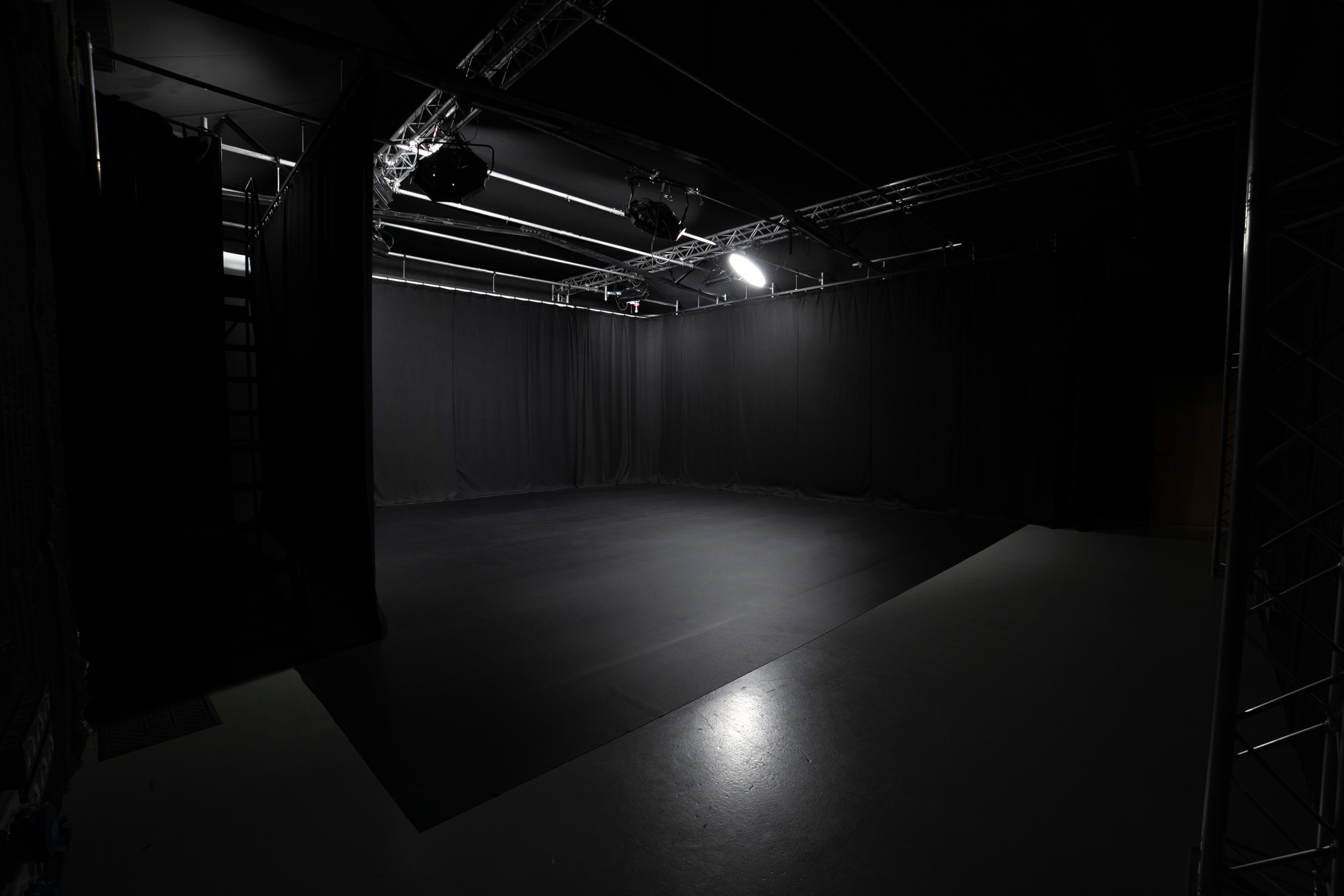The image depicts a dimly lit studio space with black curtains, empty floor, and overhead lighting.