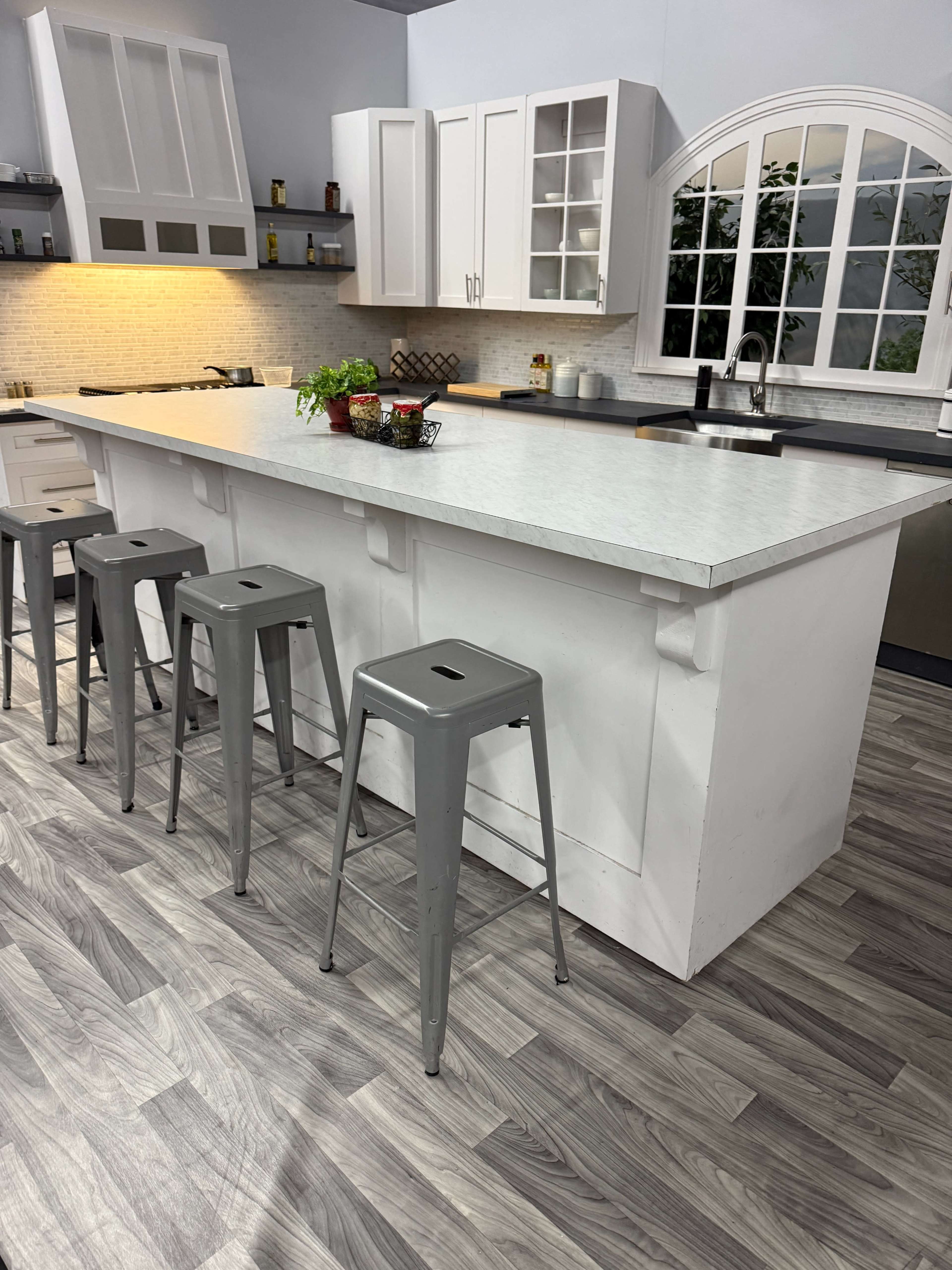 A modern kitchen features a white island with four gray metal stools on a wood-patterned floor.