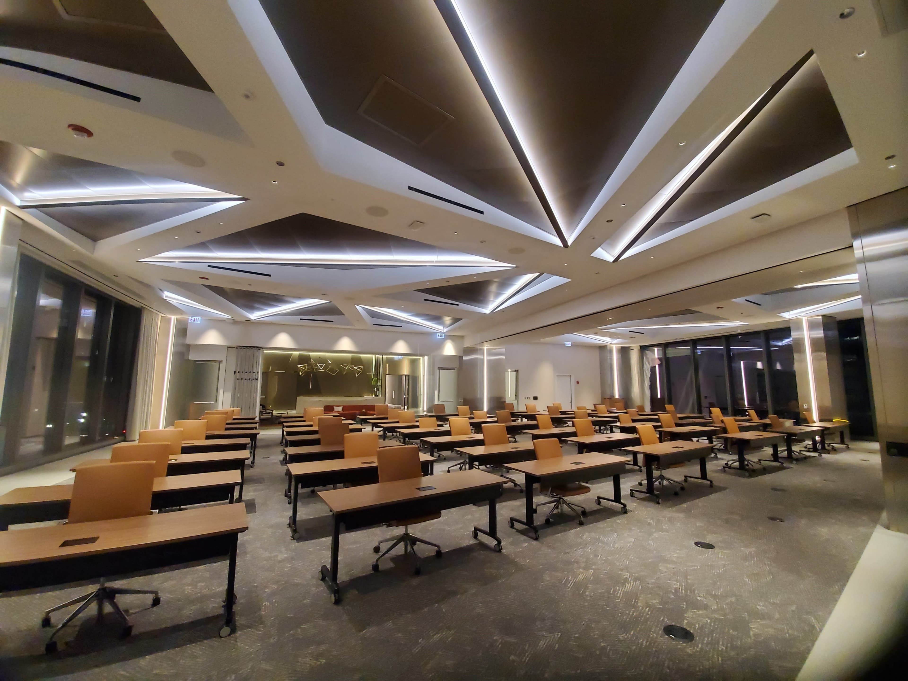 The image shows a modern, empty conference room with rows of wooden desks arranged in a spacious layout beneath a geometric ceiling with integrated lighting.
