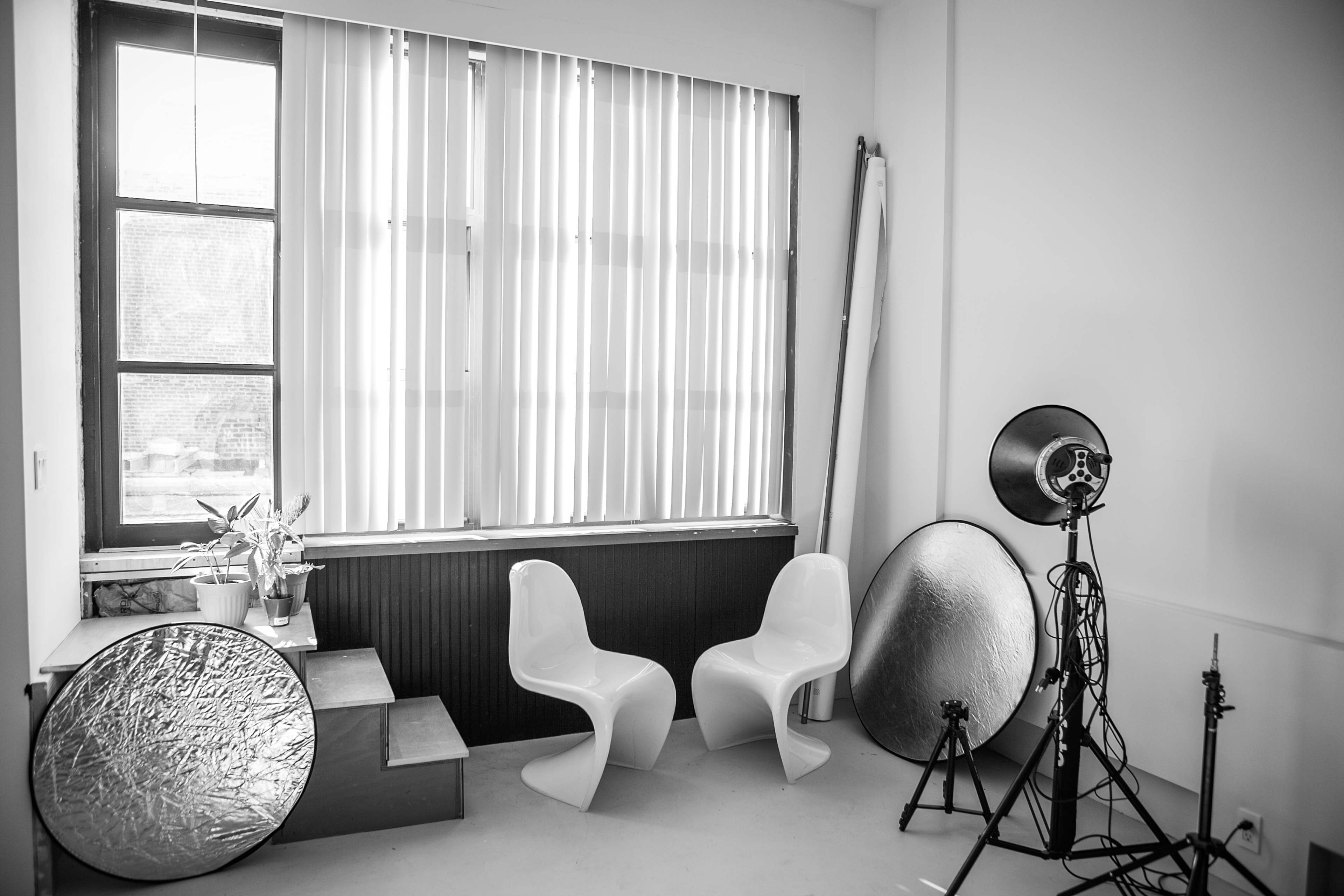 The scene shows a well-lit interior featuring two modern chairs, potted plants, and photography equipment including reflectors and a light stand.