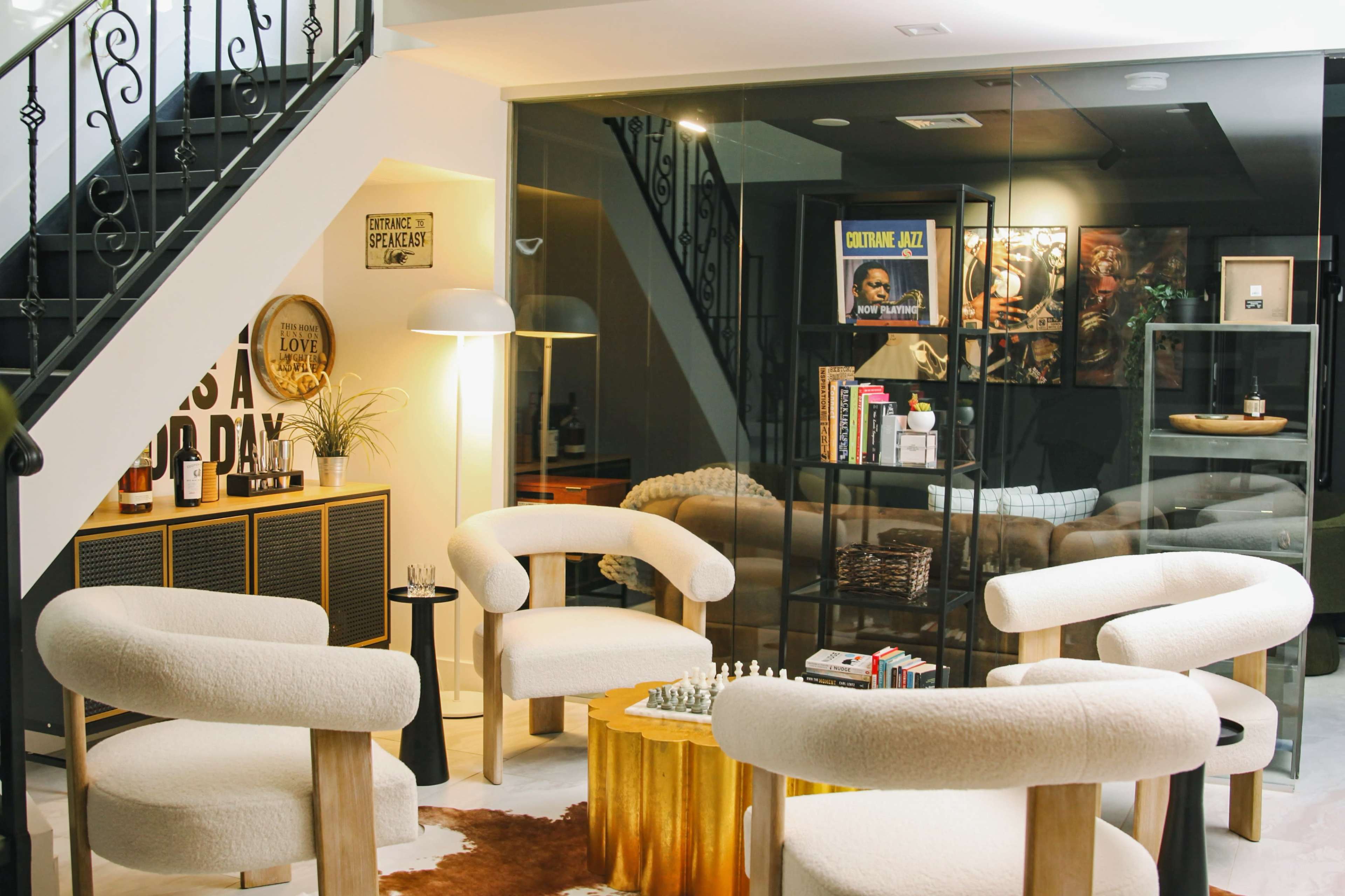 The image features a modern living area with a glass partition, stylish chairs, a small chess set on the table, and a staircase in the background.