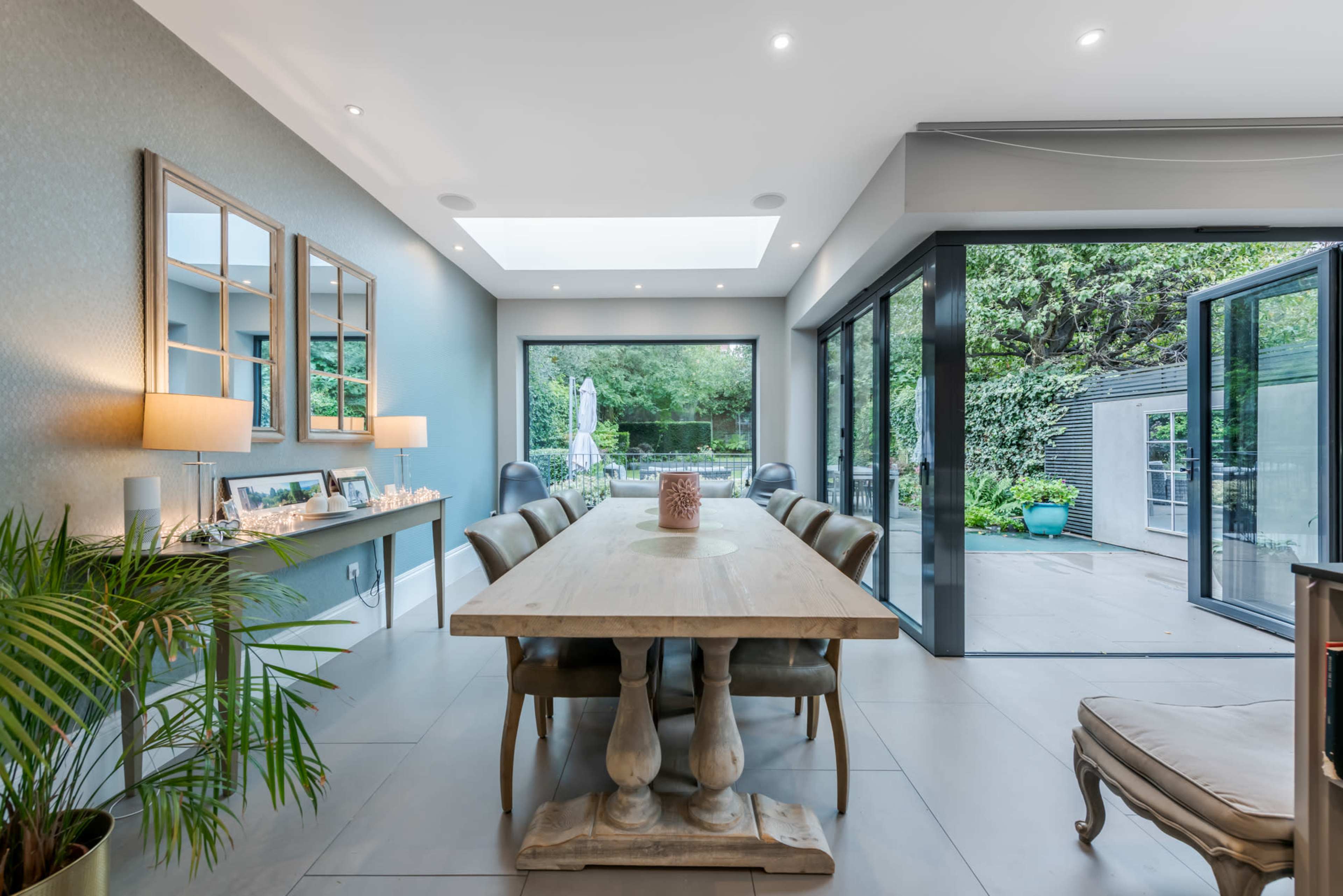 A spacious dining room features a large wooden table surrounded by upholstered chairs, with floor-to-ceiling glass doors leading to a garden.