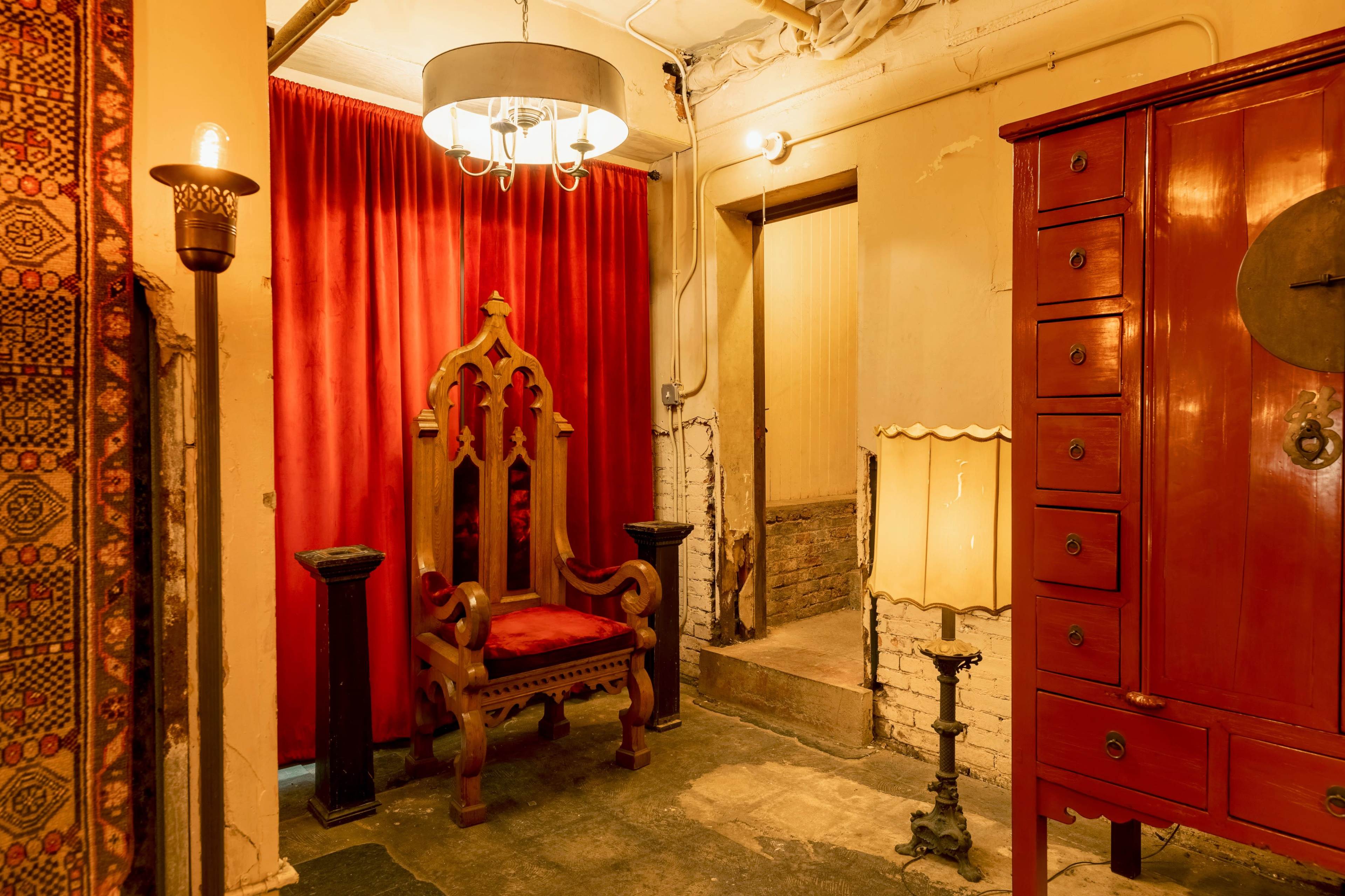 The scene features a wooden throne chair in front of a red curtain, with a red cabinet, a lamp, and an antique light fixture in a dimly lit hallway.