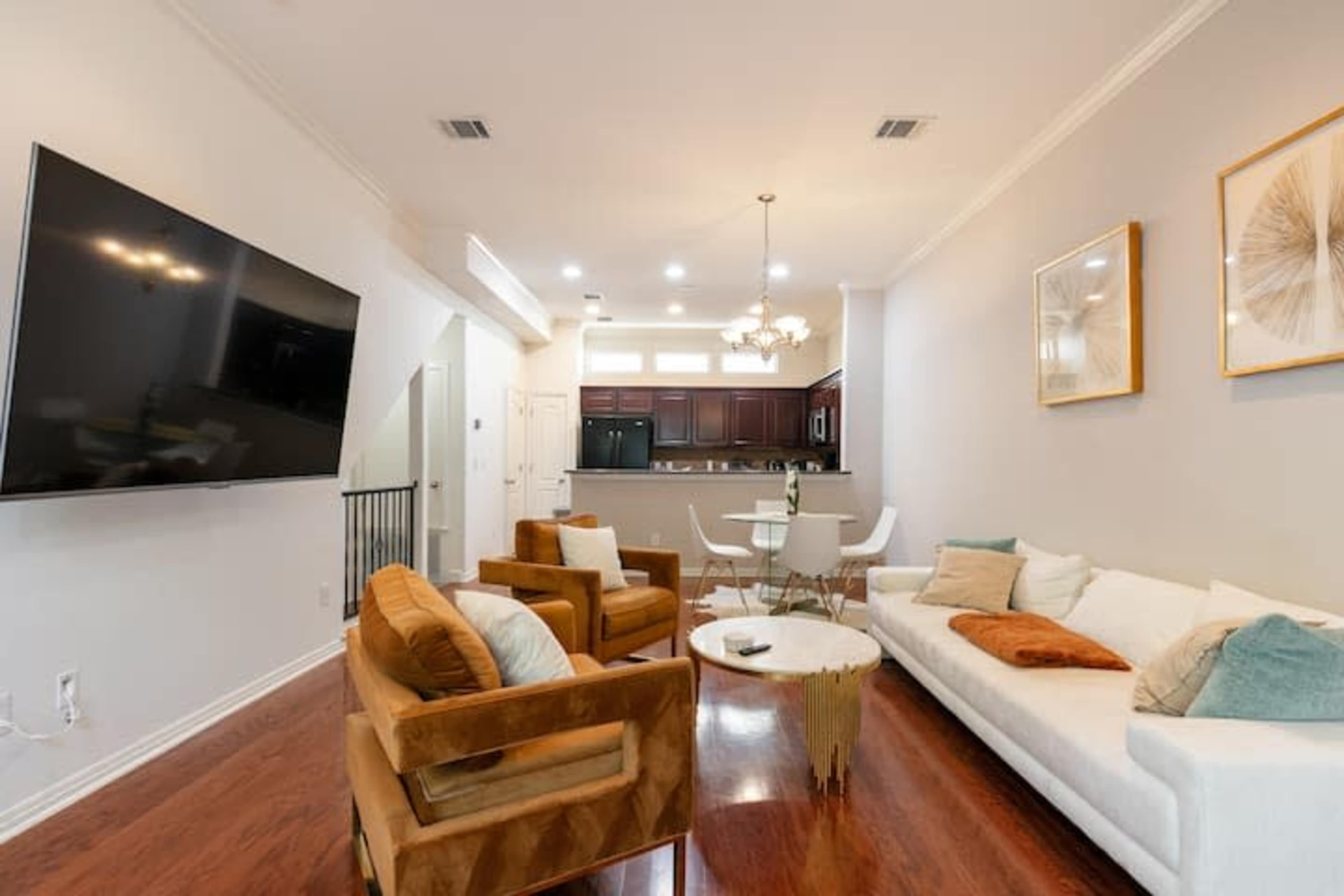 The image shows a living room with a large television mounted on the wall, a white sectional sofa, two brown armchairs, a circular coffee table, and a view into a kitchen area with dark wood cabinets.