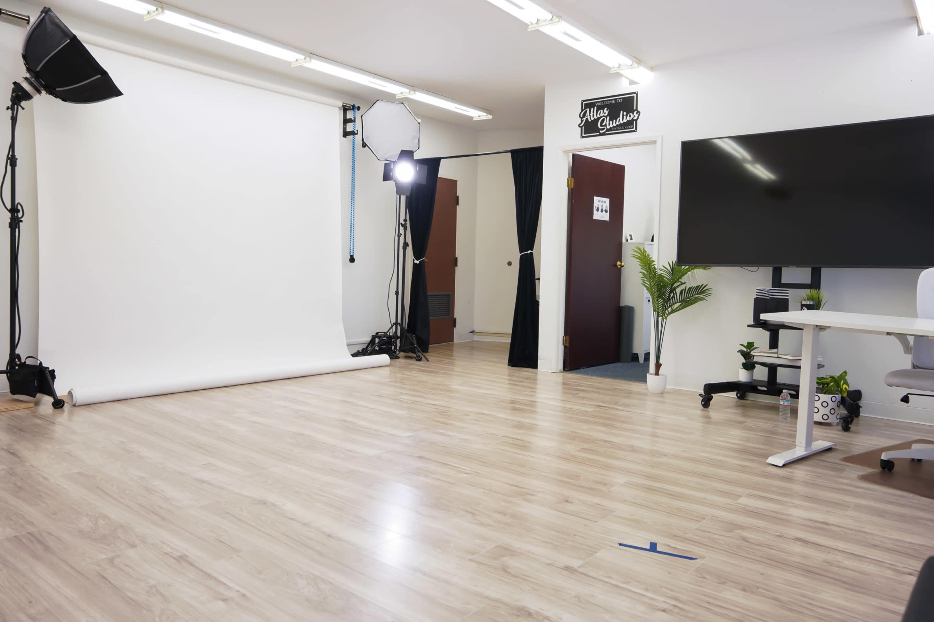 A well-lit studio with a white backdrop, softbox lights, and a desk with a monitor in a minimally decorated space.