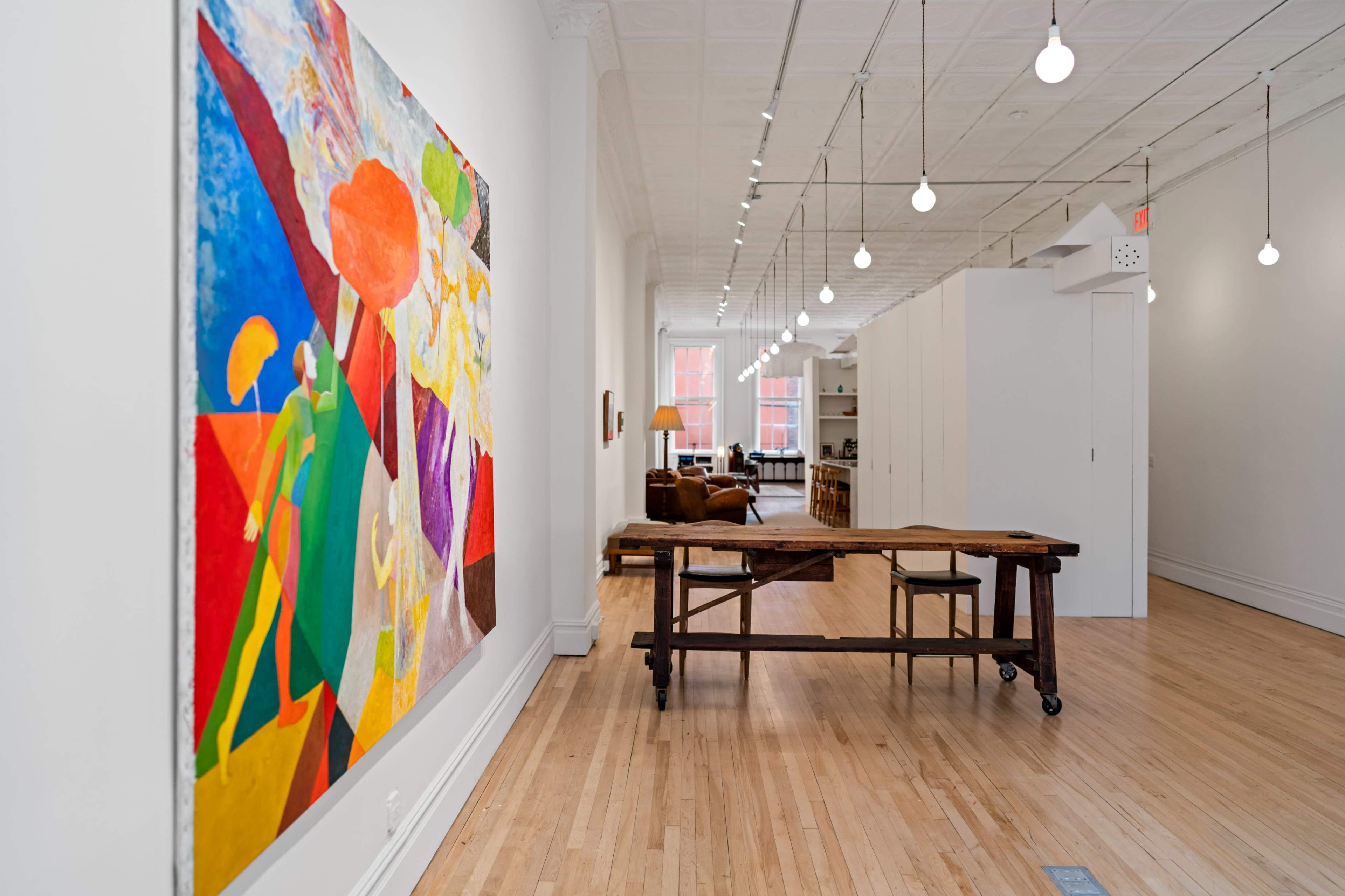 A brightly lit gallery space featuring a large colorful painting on the left wall and a wooden table with two stools in the center, leading to a cozy living area in the background.
