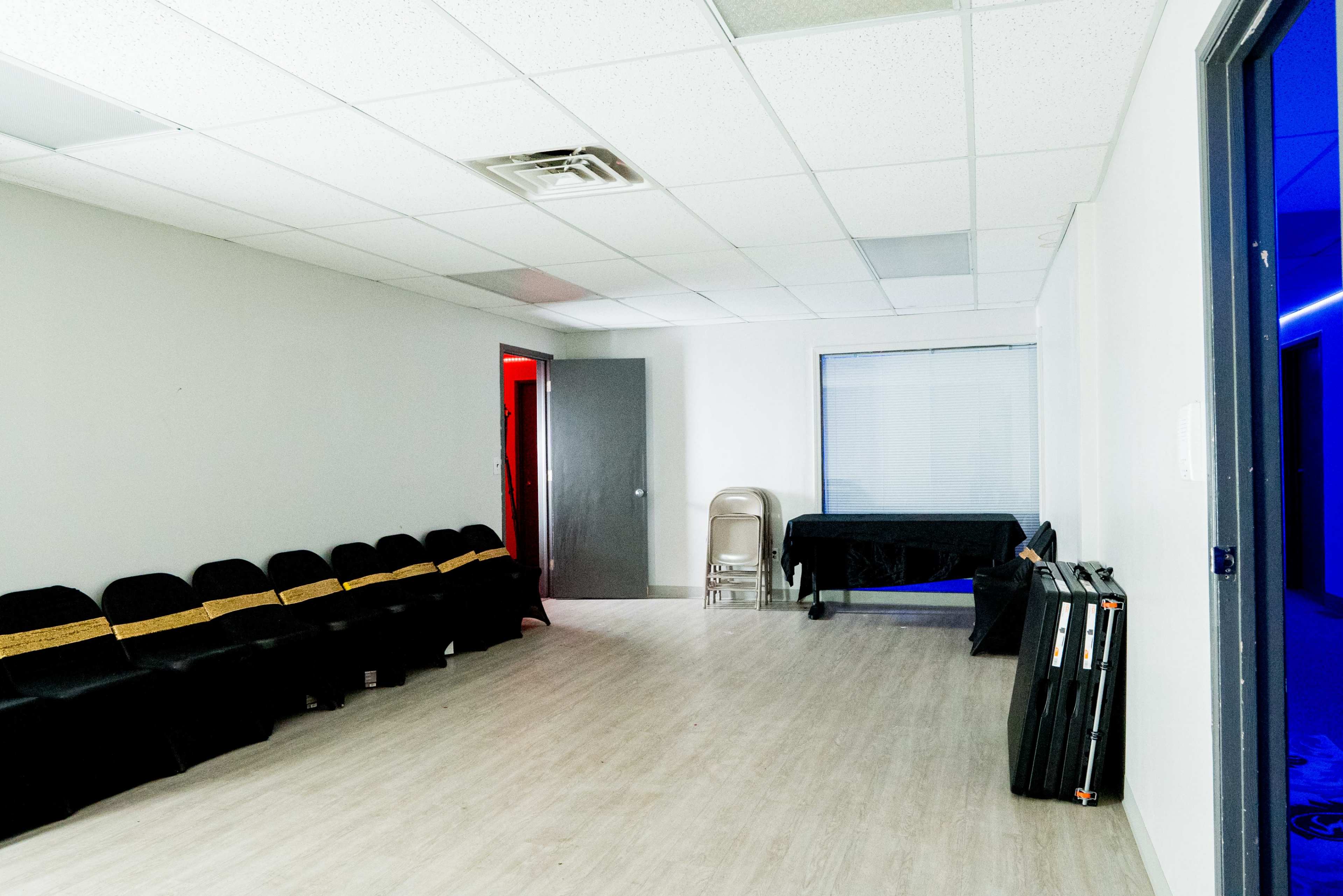 The image shows a sparsely furnished room with black chairs arranged in a row, a folding chair, a table covered with a dark cloth, and a blue-lit window.
