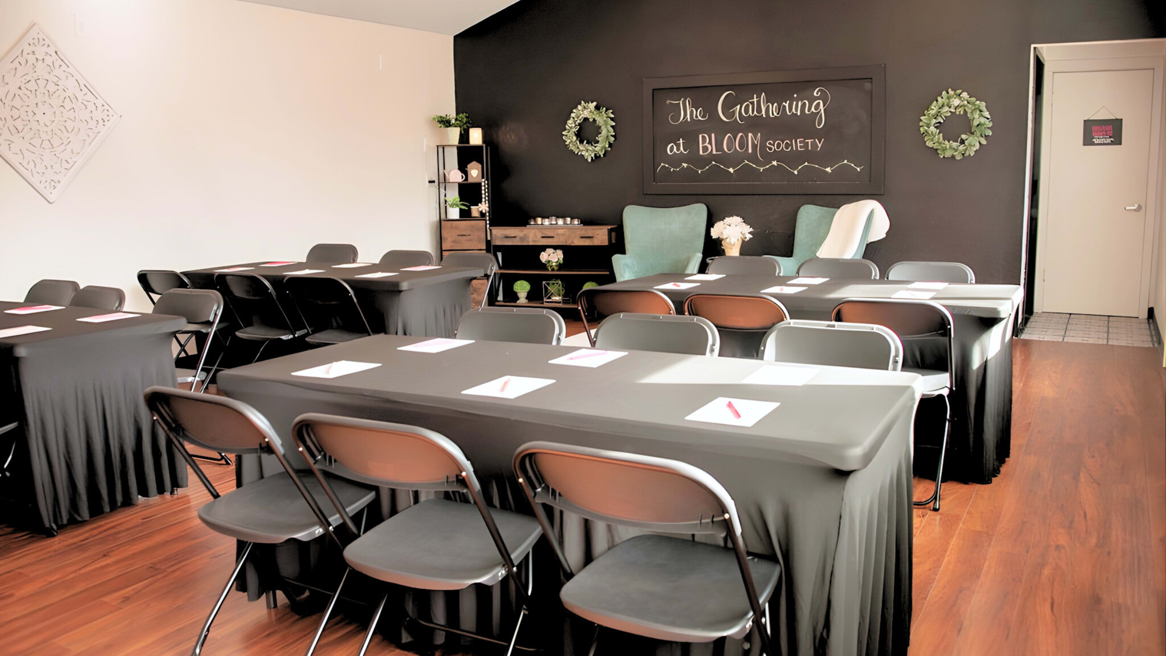 The image shows a classroom setup with black tables and chairs arranged in a u-shape, featuring a dark wall with decorative elements and a sign that reads "The Gathering at BLOCC Society."