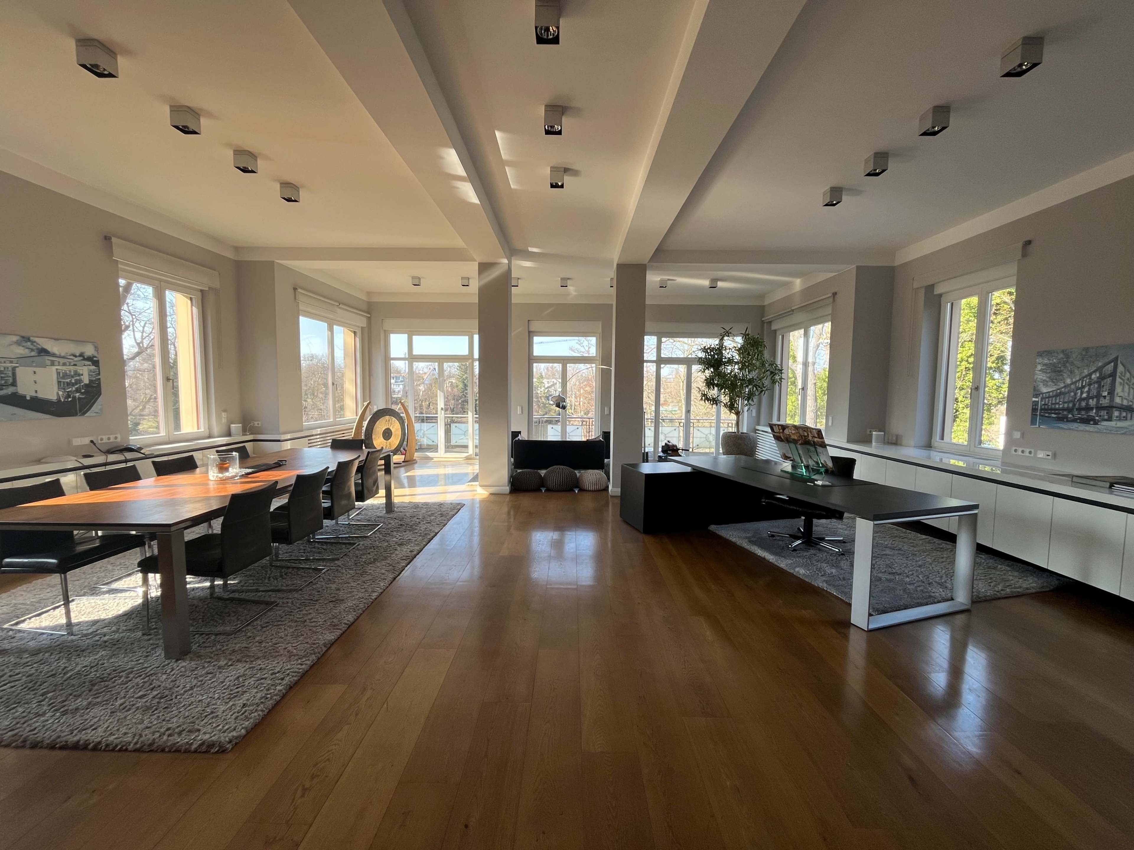 The image shows a spacious, modern living area with a dining table, chairs, and large windows that provide a view of the outdoors.