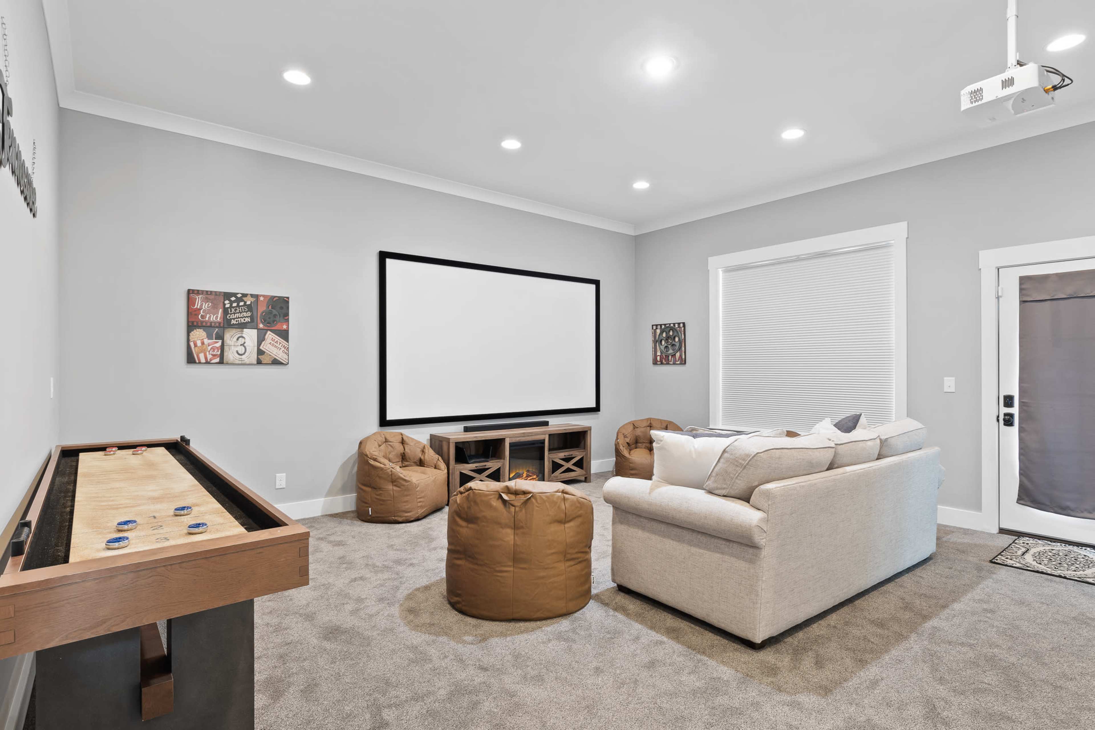 A modern, cozy entertainment room featuring a large screen, a sofa, bean bag chairs, and a shuffleboard table.
