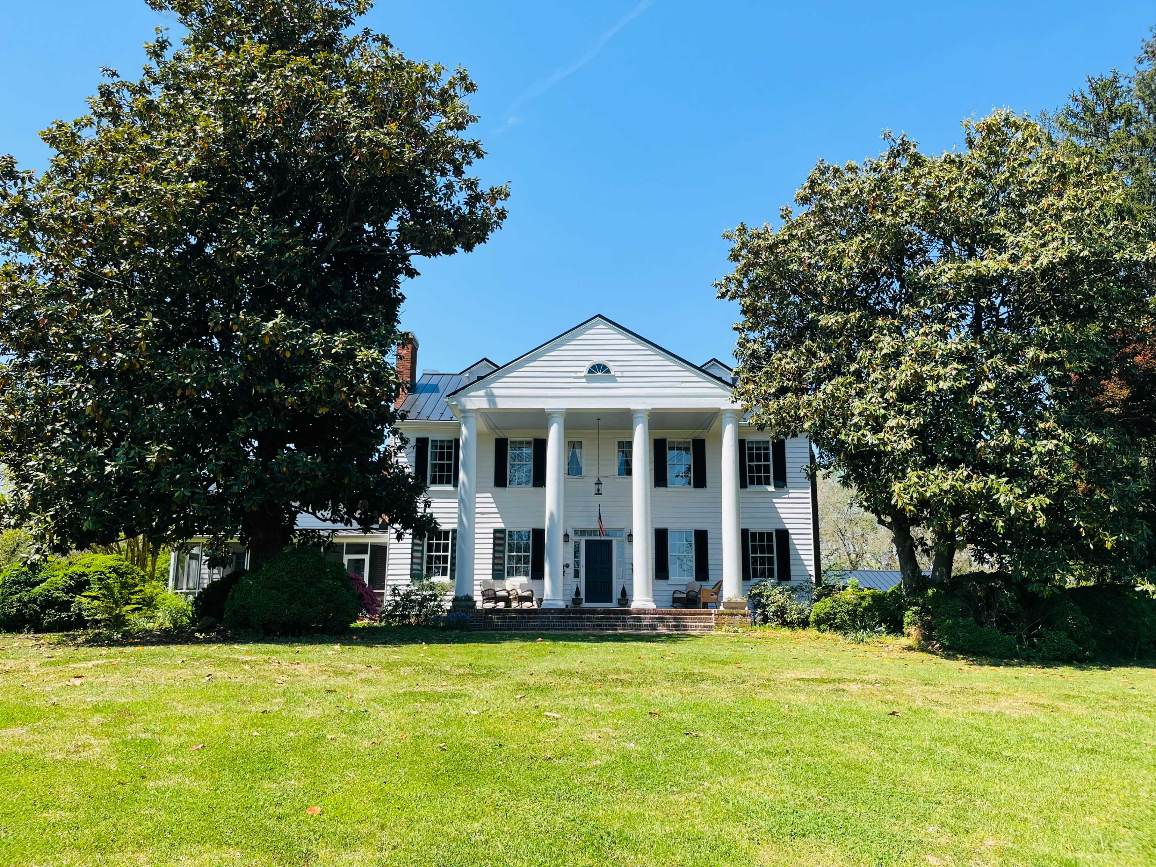 A large white colonial-style house with six columns and a manicured lawn is surrounded by tall trees under a clear blue sky.