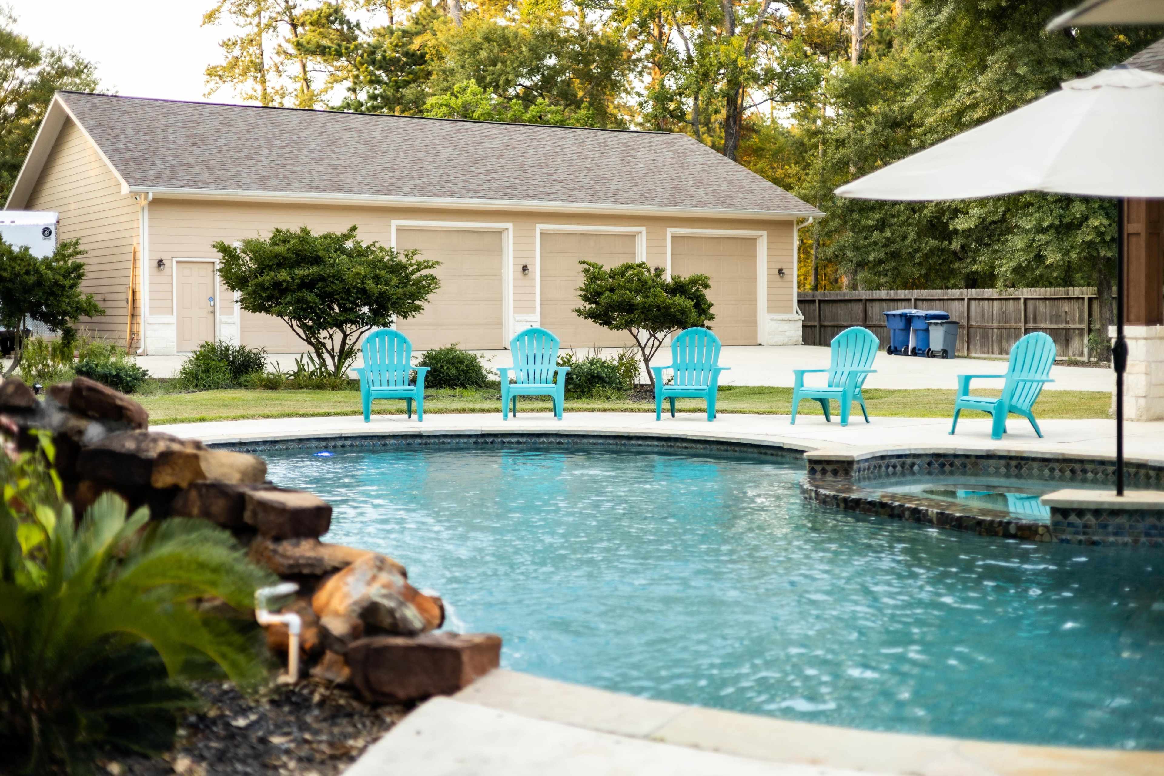 A swimming pool with a stone edge and blue chairs is situated in front of a garage with three doors in a backyard setting.