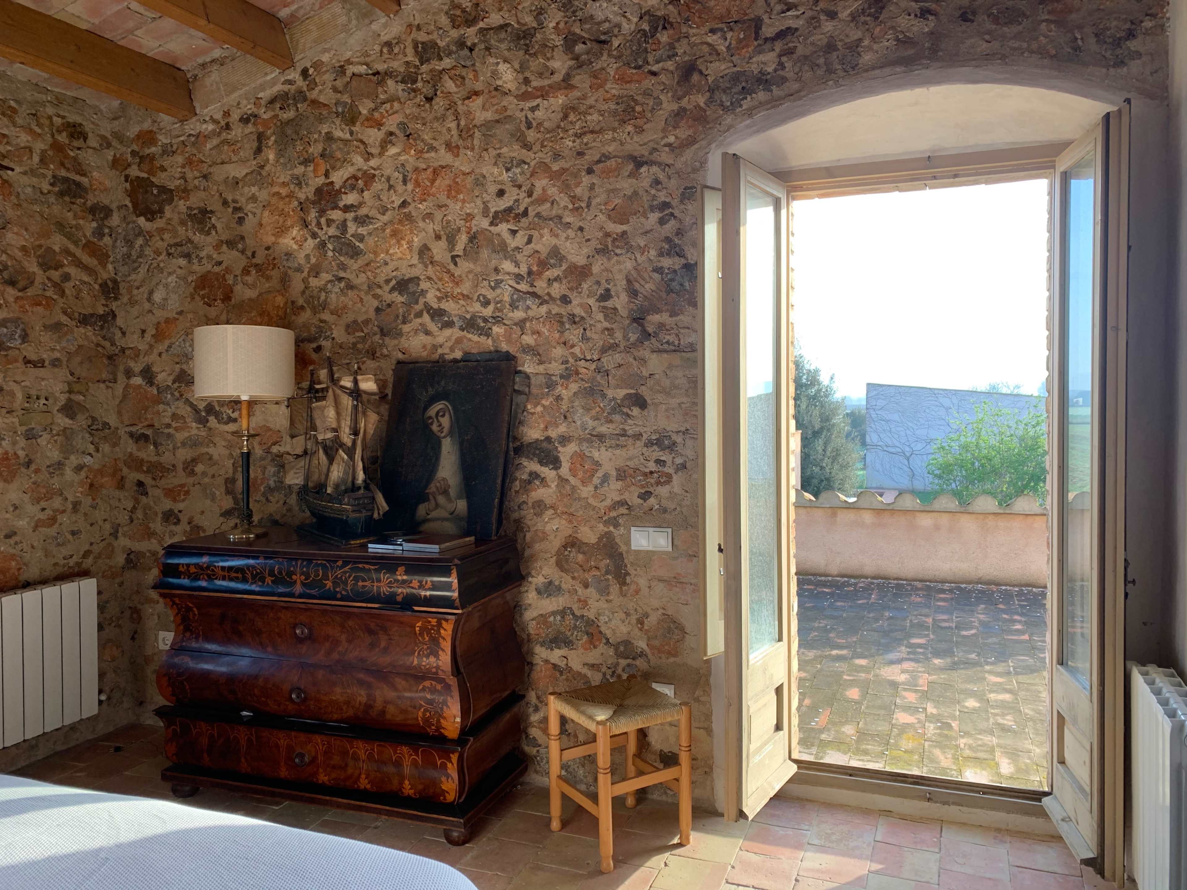 A room with exposed stone walls features a wooden dresser, a painting, and an open door leading to a terrace.