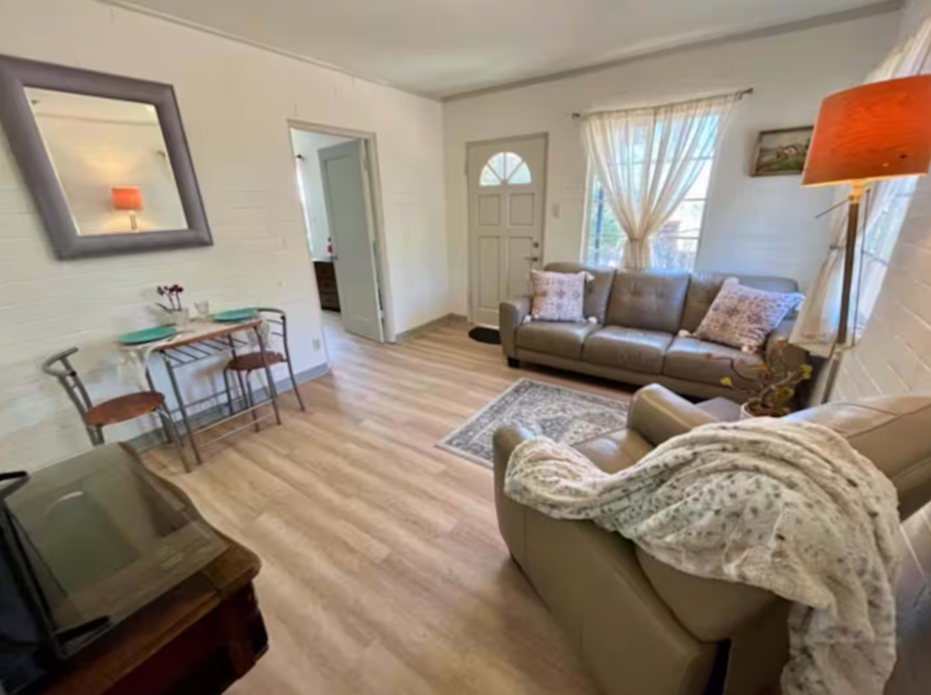 The image shows a light-filled living area with a brown leather sofa, a small dining table, and hardwood flooring.