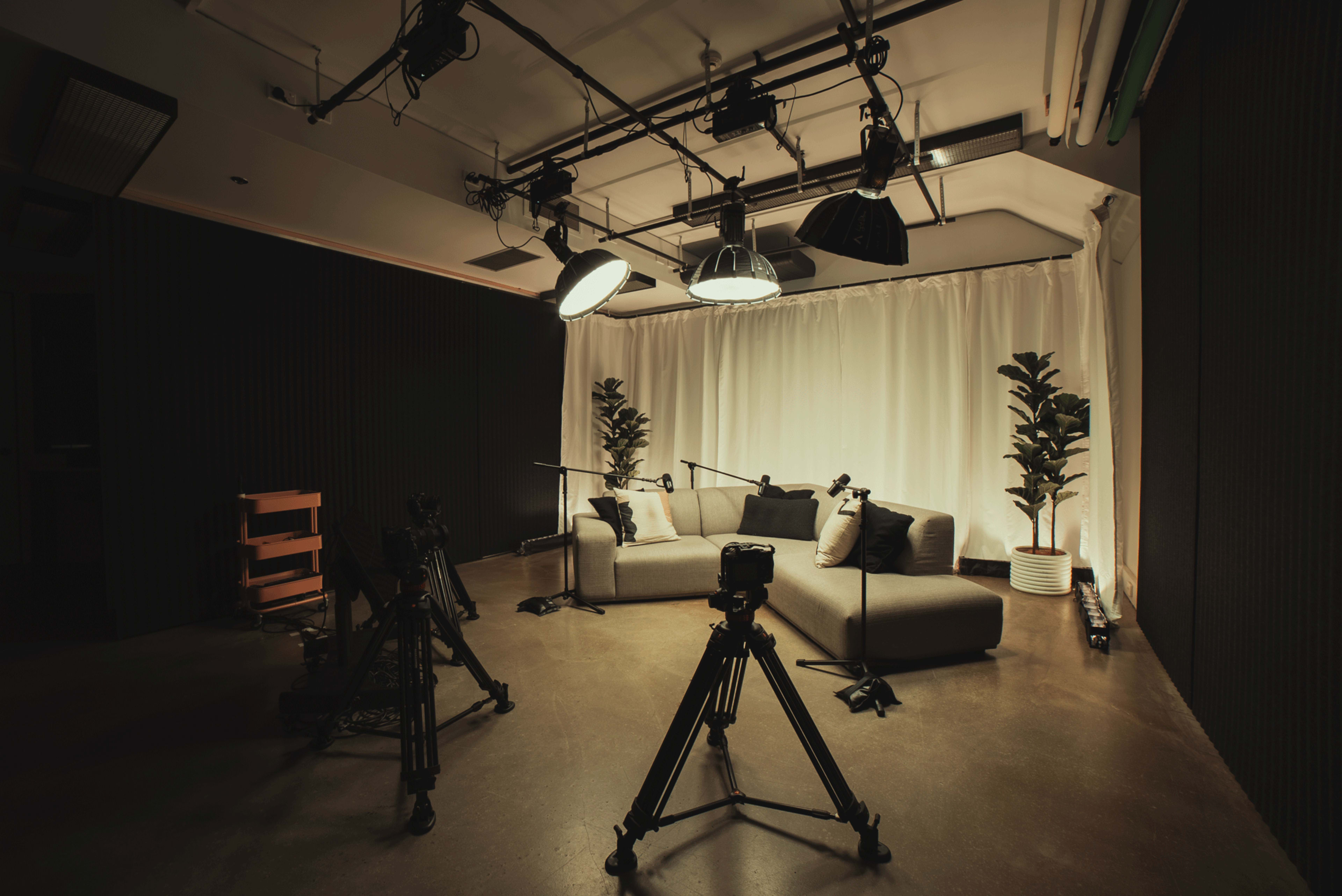 A television studio is set up with a couch, two potted plants, and multiple cameras positioned on tripods, all under bright studio lights.
