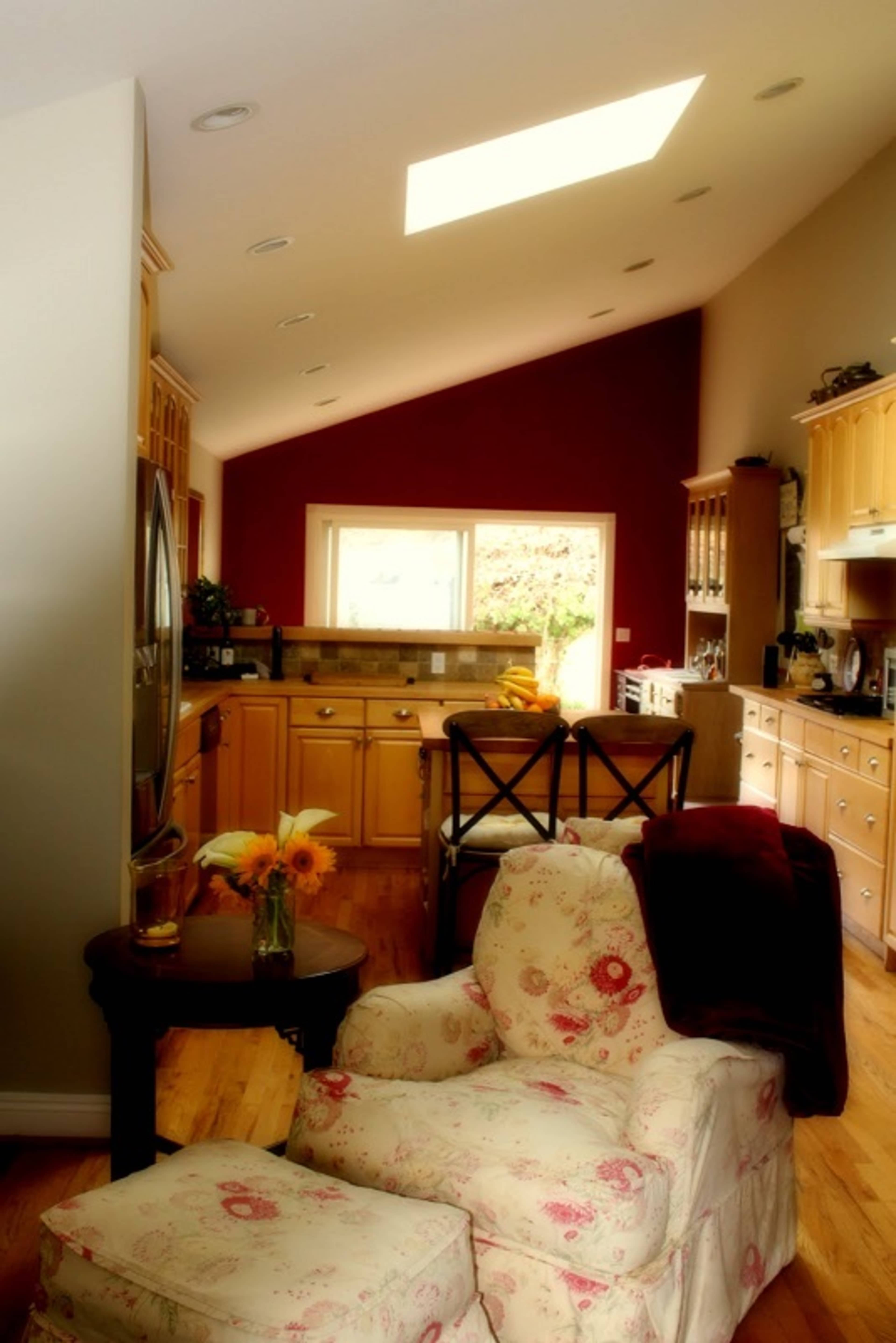 A brightly lit kitchen features wooden cabinetry, a skylight, and a floral-patterned armchair with a small table and fresh flowers.