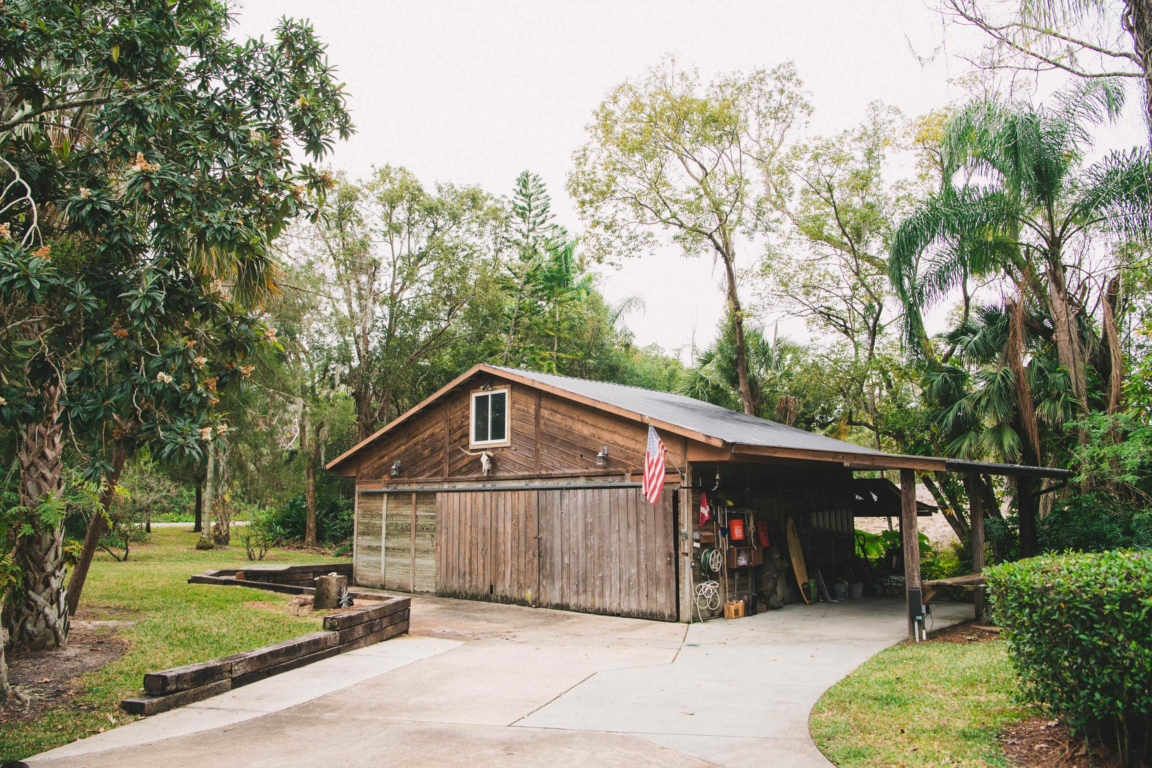 A wooden house with a covered porch is surrounded by trees and greenery in a rural setting.