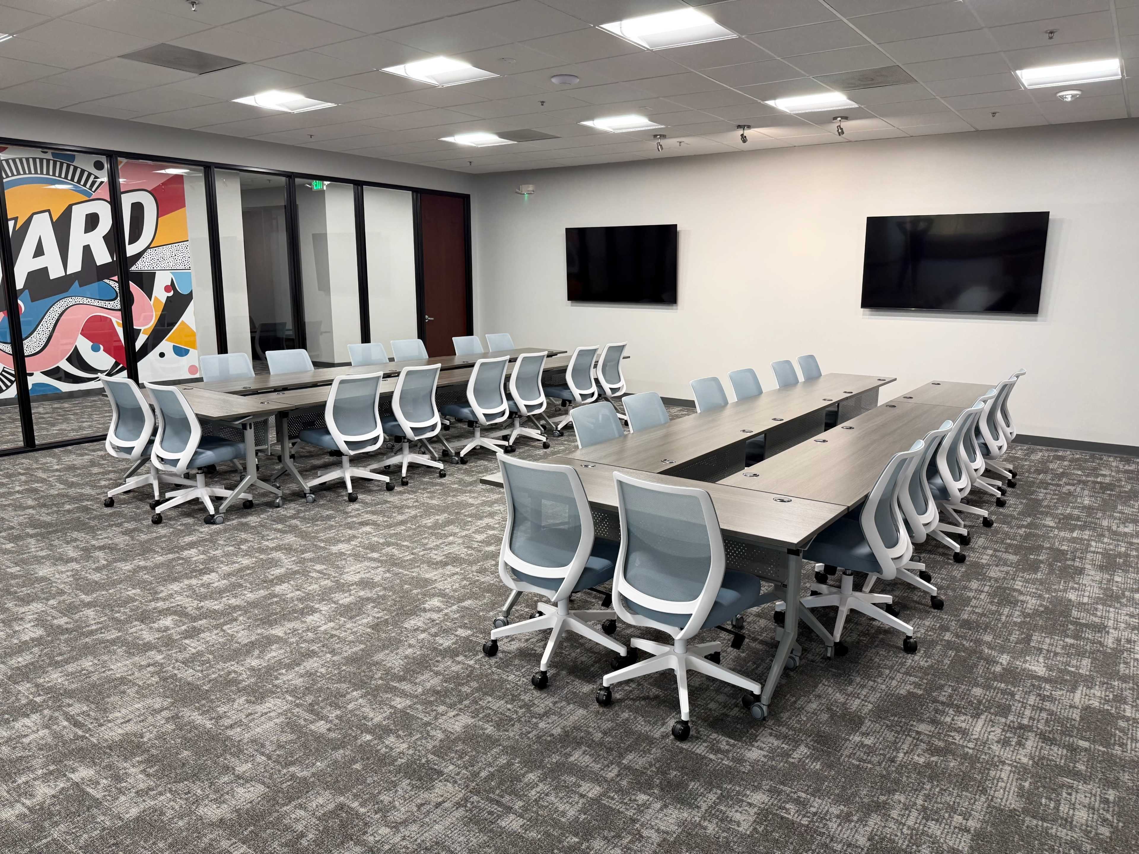 A modern conference room features a long table with multiple seating arrangements and two large screens mounted on the walls.
