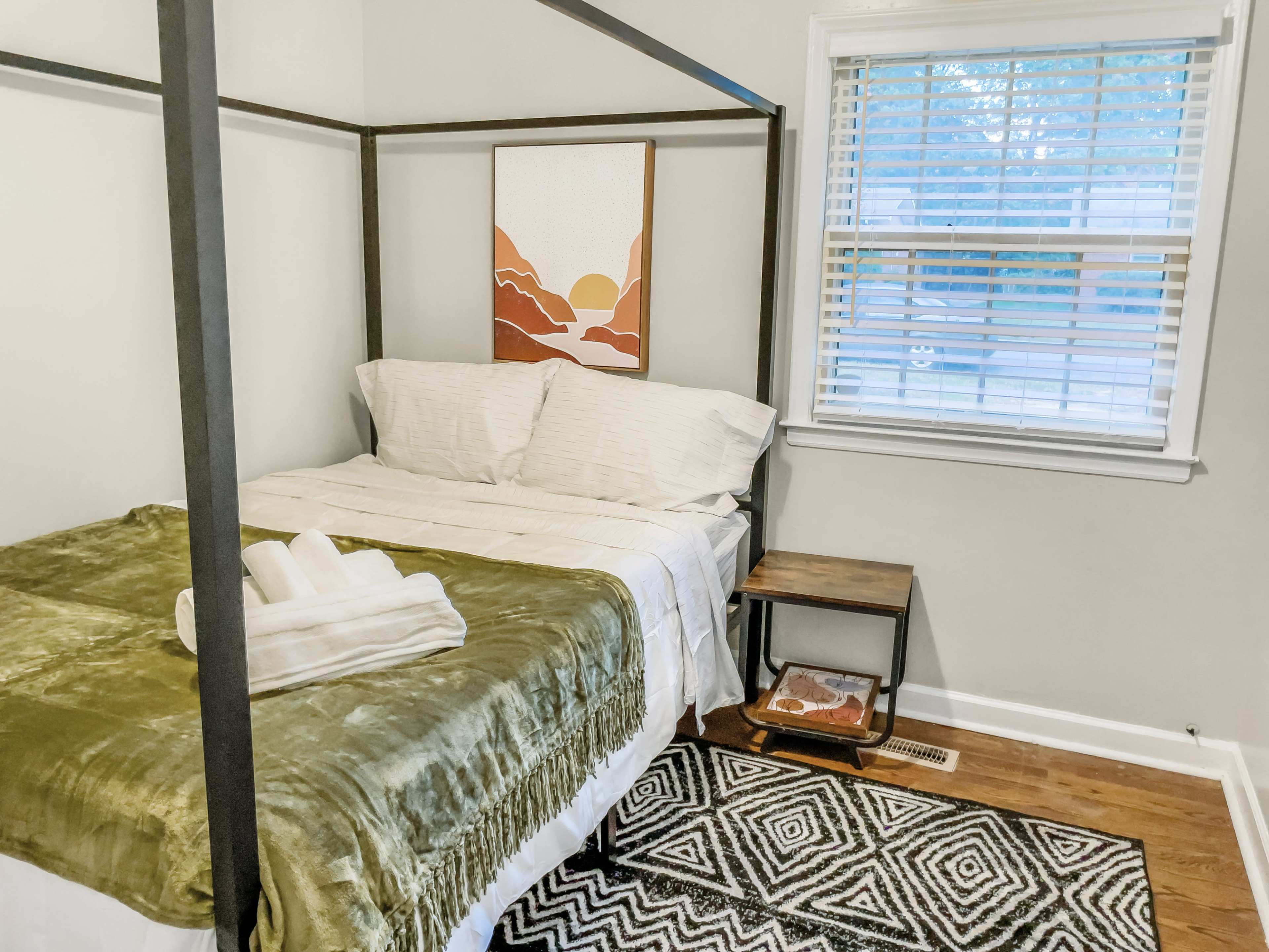 A canopy bed with white bedding and a green throw, accompanied by a nightstand and a framed artwork on the wall, in a well-lit bedroom with a window.