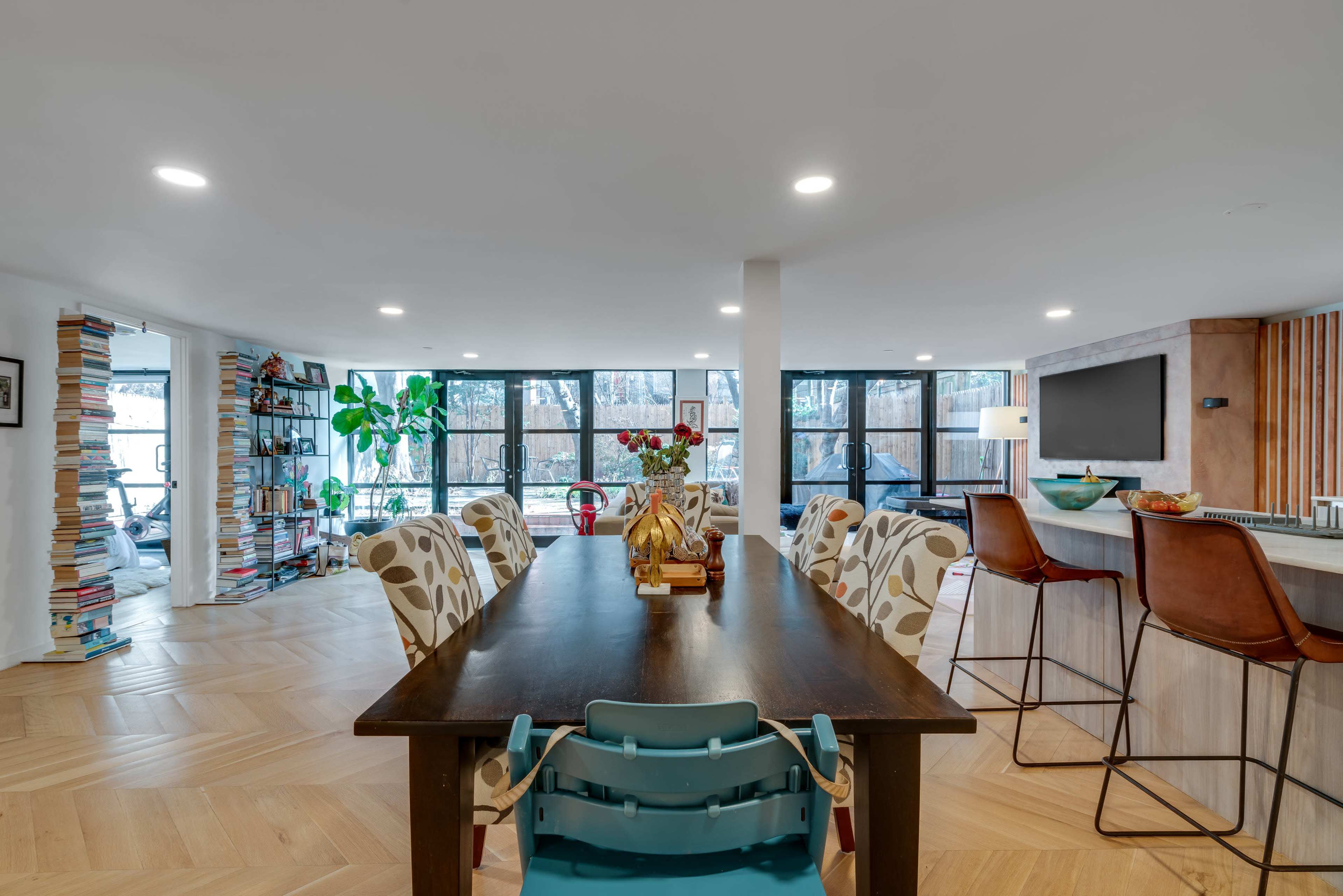 A modern dining area features a long table surrounded by patterned chairs, with large windows allowing natural light in and bookshelves lining one wall.
