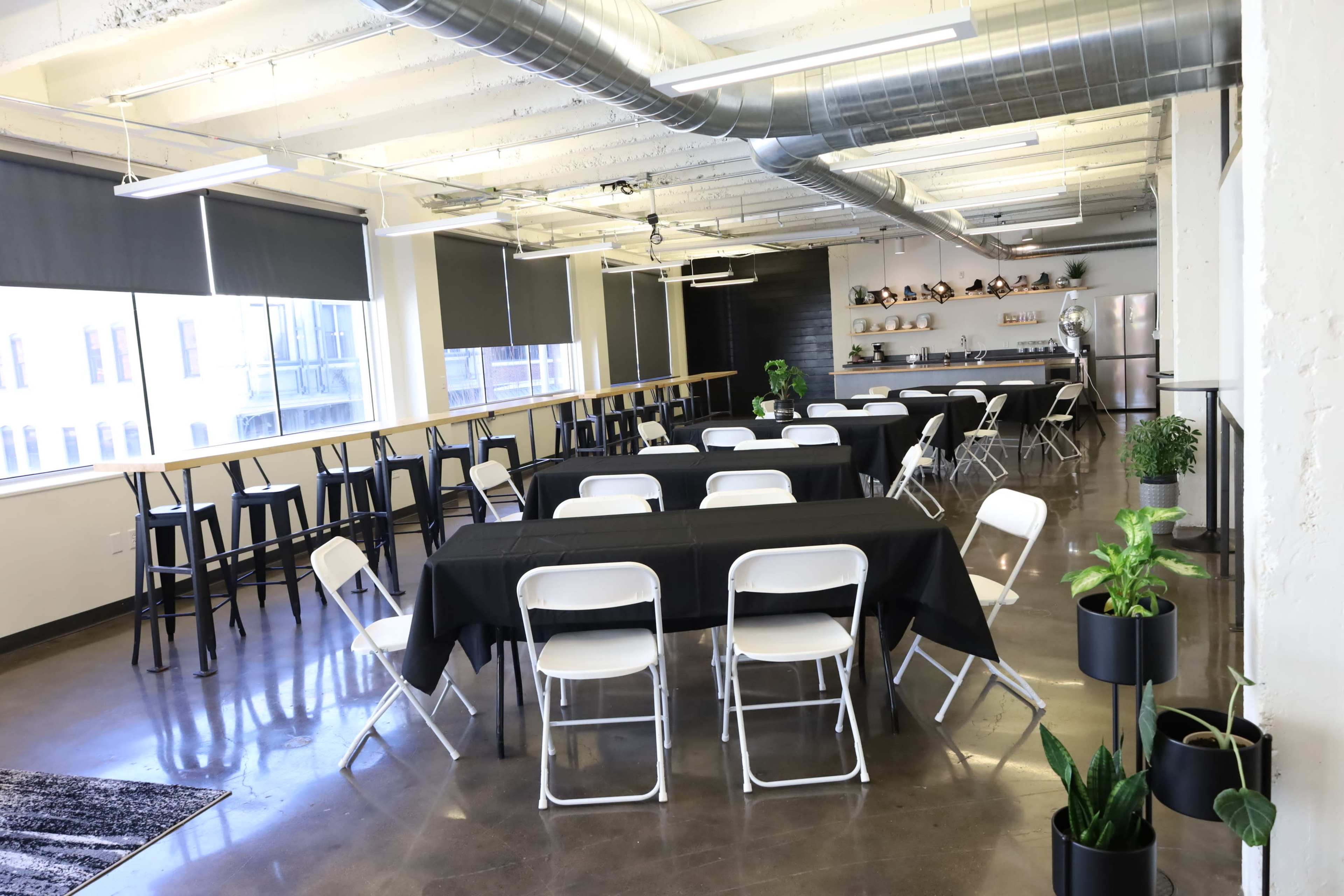 A brightly lit event space with black tables, white folding chairs, and plants, featuring large windows and a kitchenette area in the background.