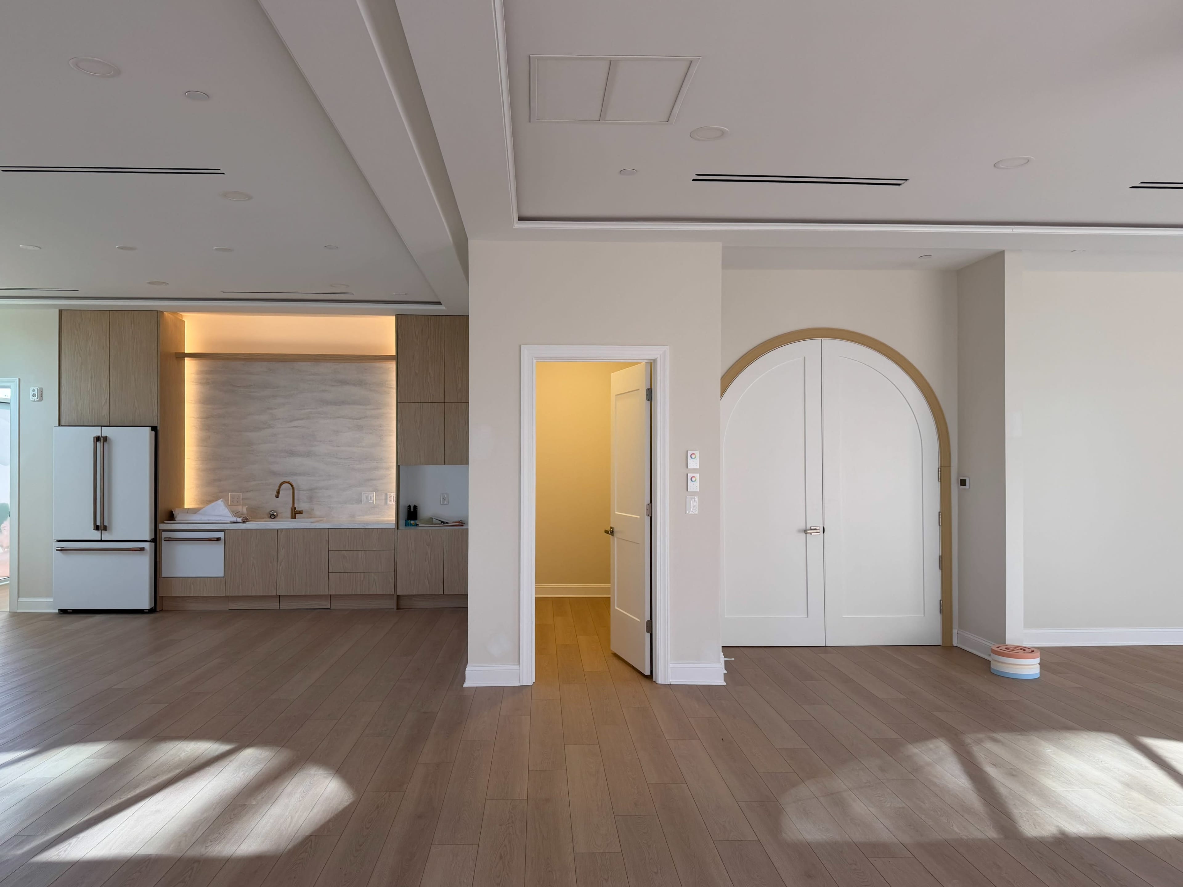 The image shows an open-concept living space featuring a kitchen area with wood cabinetry and a white refrigerator, alongside a large arched doorway leading to a separate room.