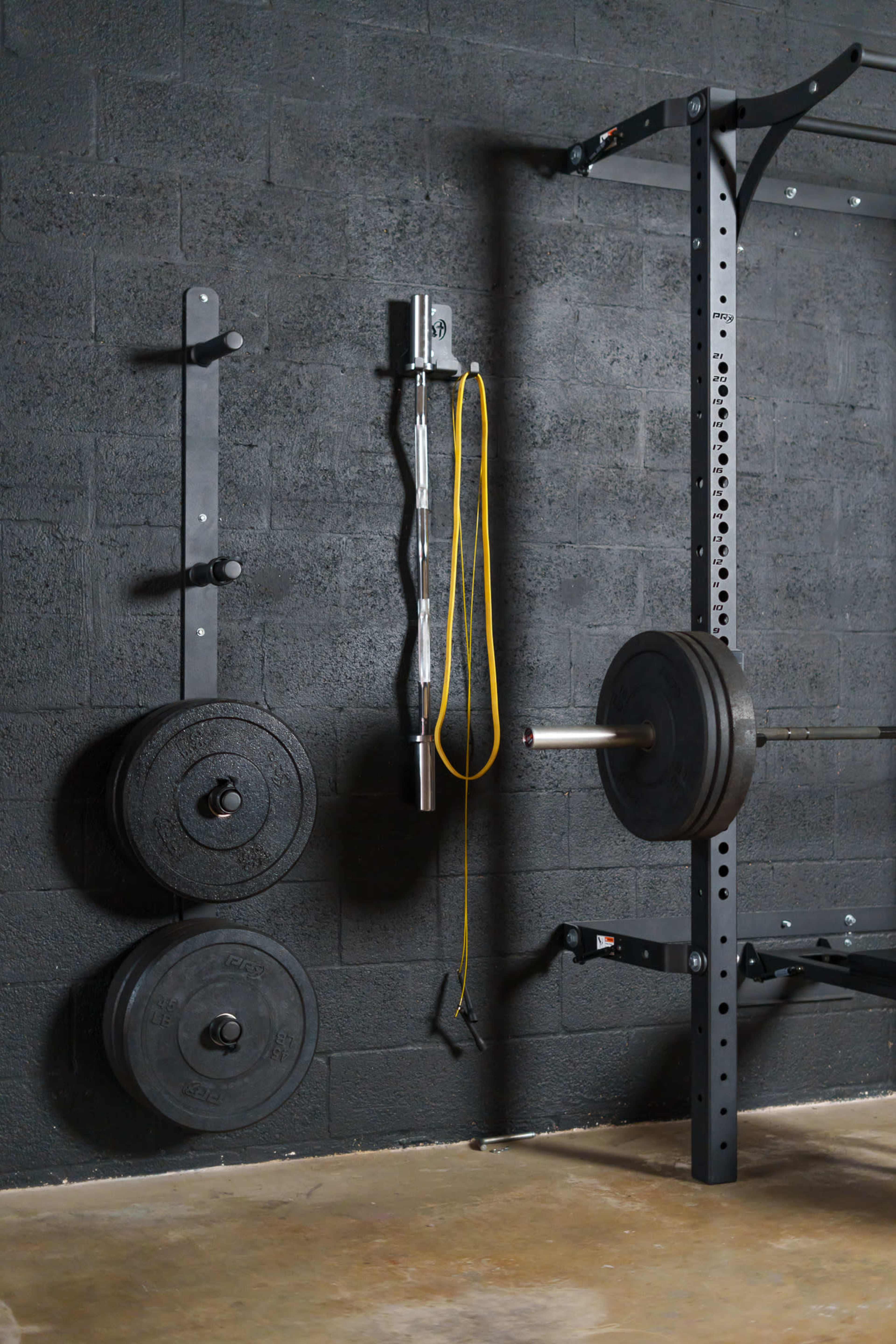 A weightlifting setup with a barbell mounted on a rack, weight plates stored on a wall, and a jump rope hanging next to it against a dark concrete wall.