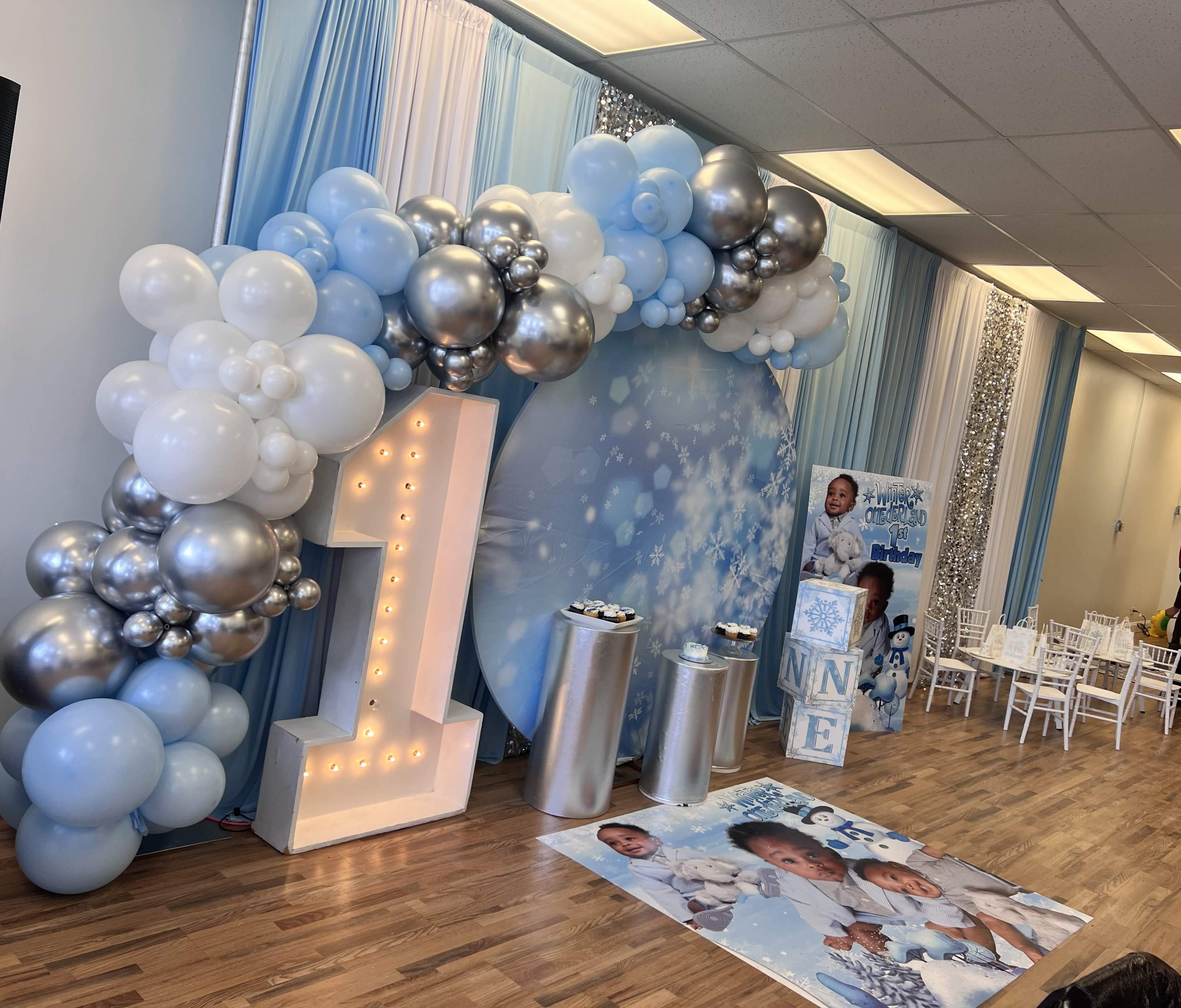 The scene features a birthday party setup with a large illuminated number "1" surrounded by blue and silver balloons, a decorative backdrop, and tables arranged for guests.