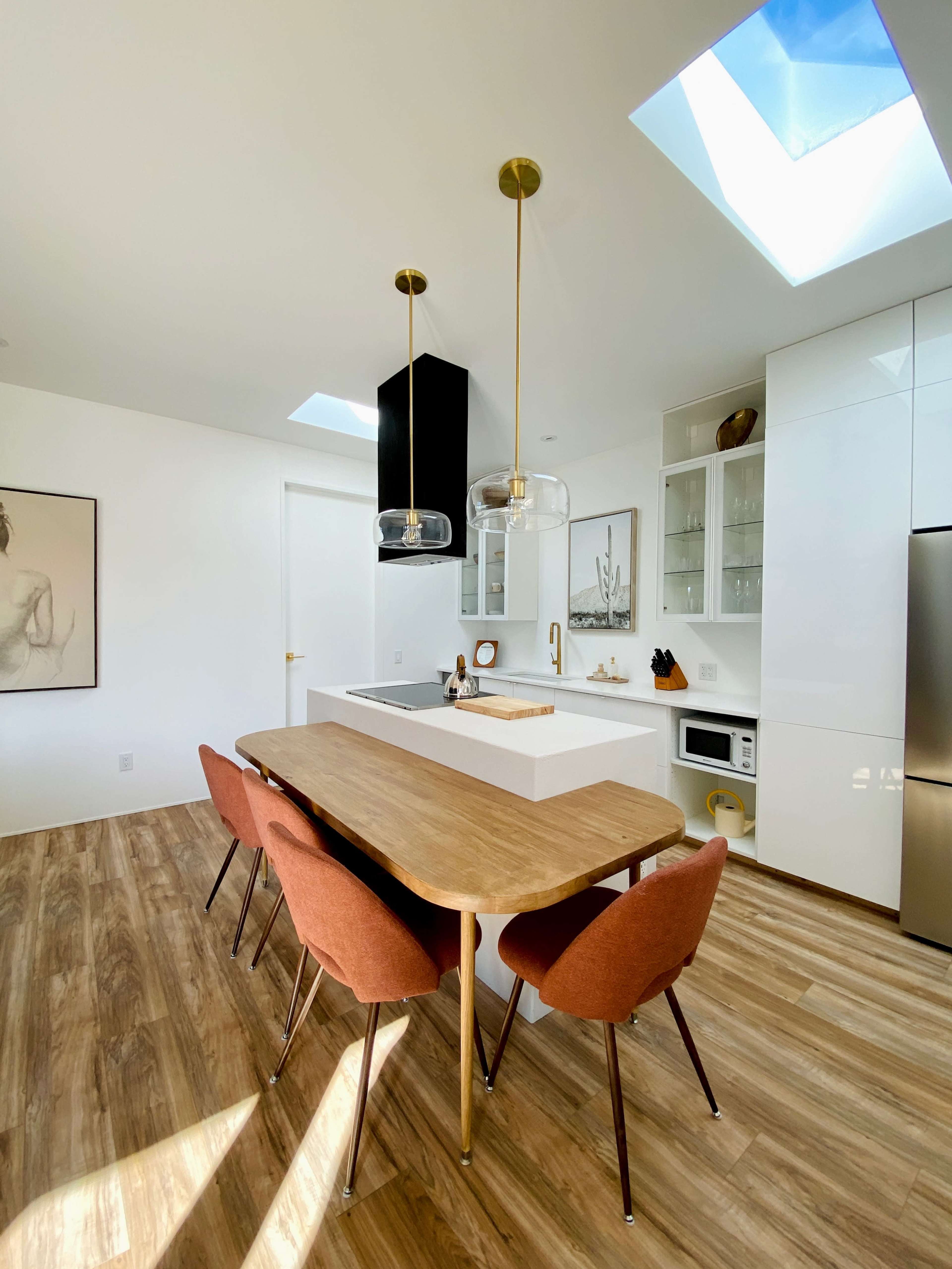 A modern kitchen features a central wooden dining table surrounded by brown chairs, with sleek cabinetry and pendant lights above.