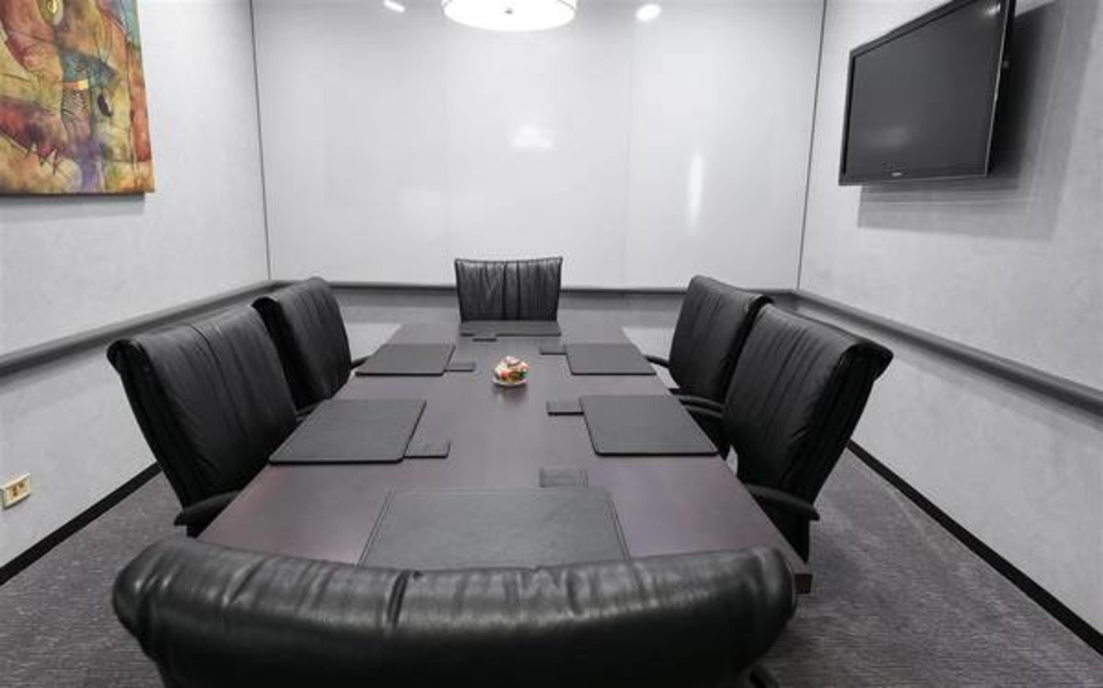 A rectangular conference room table surrounded by eight black leather chairs is positioned under a circular light fixture, with a wall-mounted television and a piece of art hanging nearby.