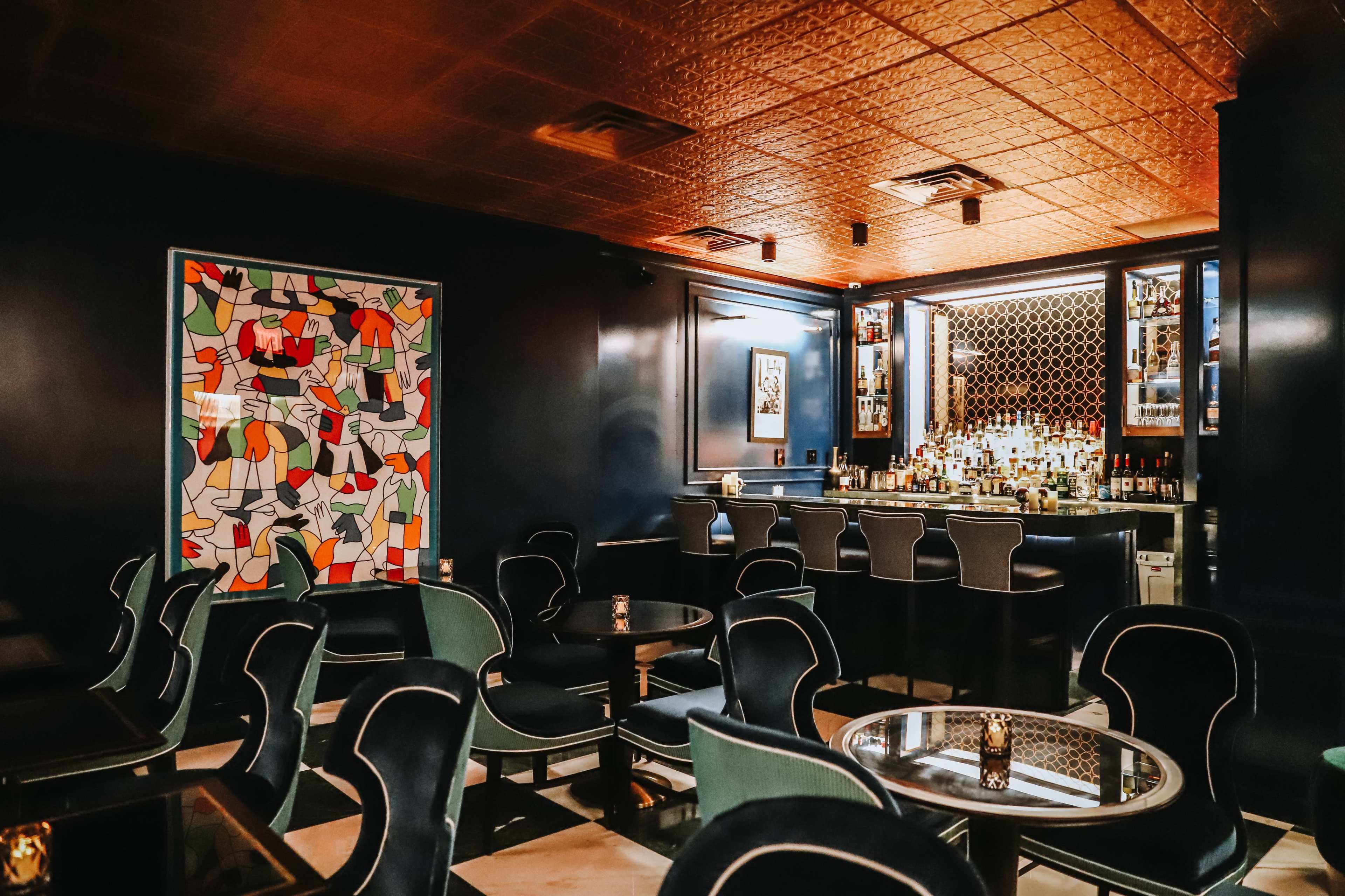 A dimly lit bar area with modern seating, a patterned artwork on the wall, and a well-stocked bar in the background.