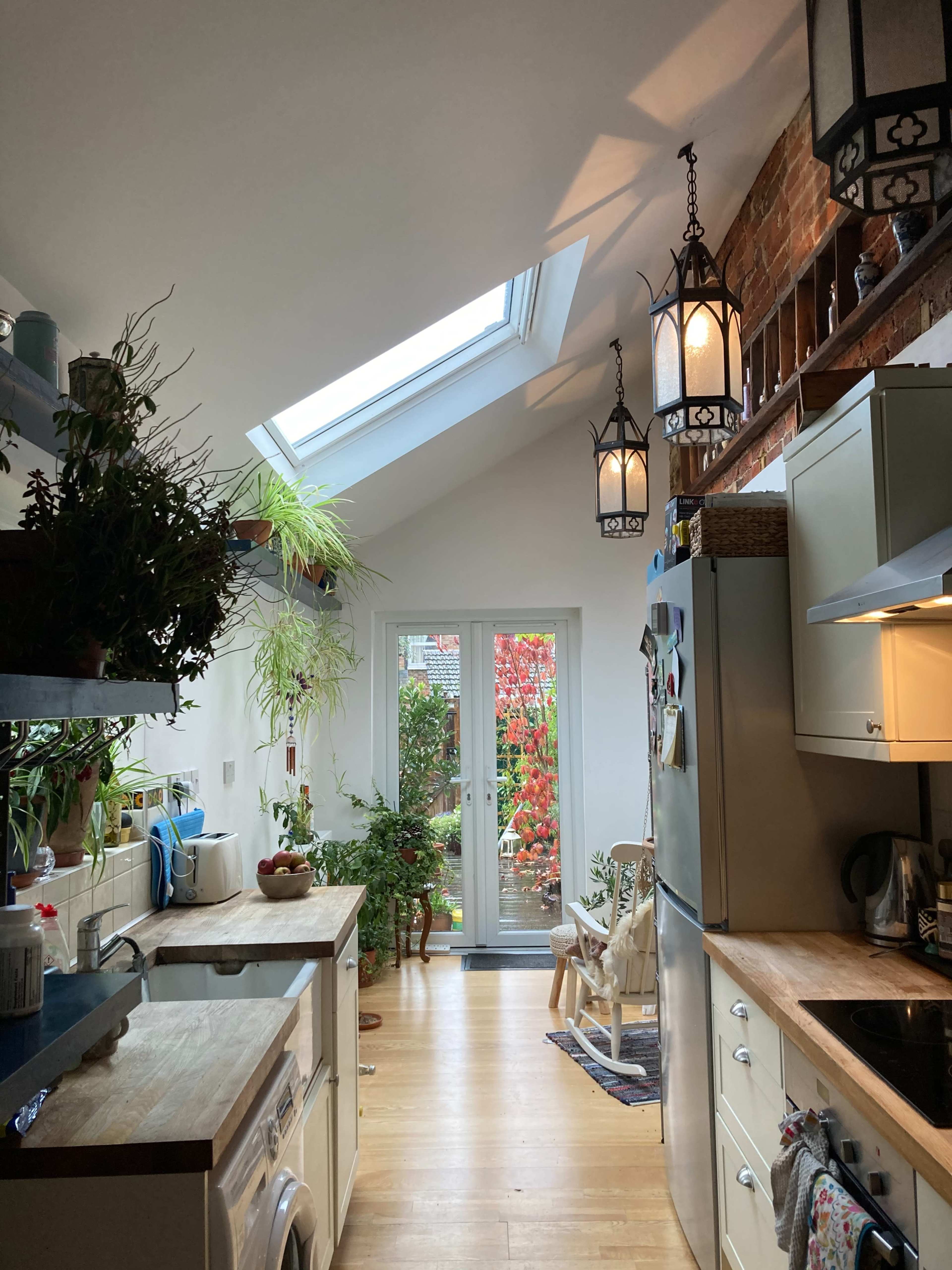A well-lit kitchen features a skylight, potted plants, and various appliances, leading to a glass door that opens to the garden.