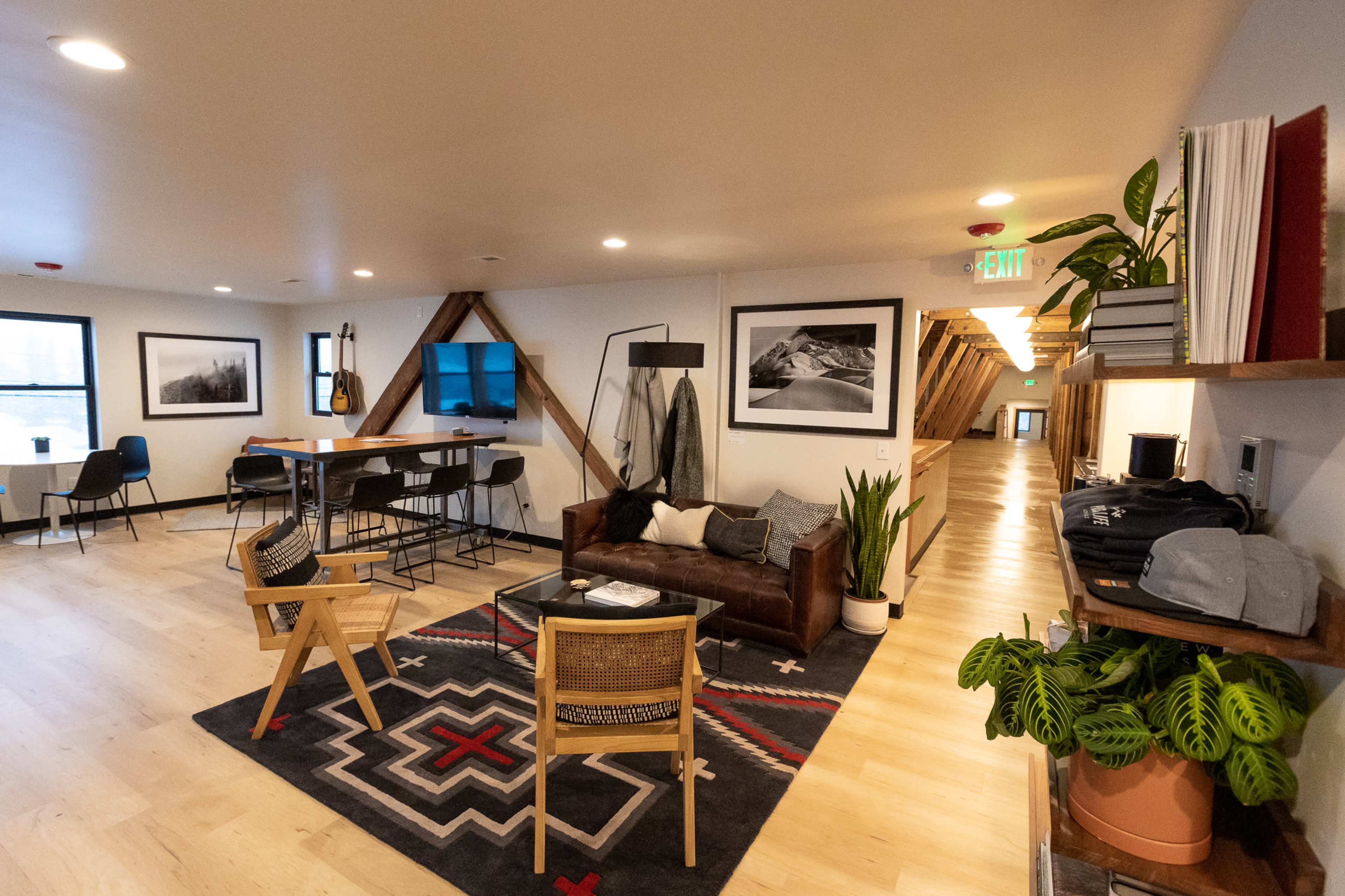 The image shows a modern open-concept lounge area featuring a leather sofa, wooden chairs, and a rug, with a dining space and hallway in the background.
