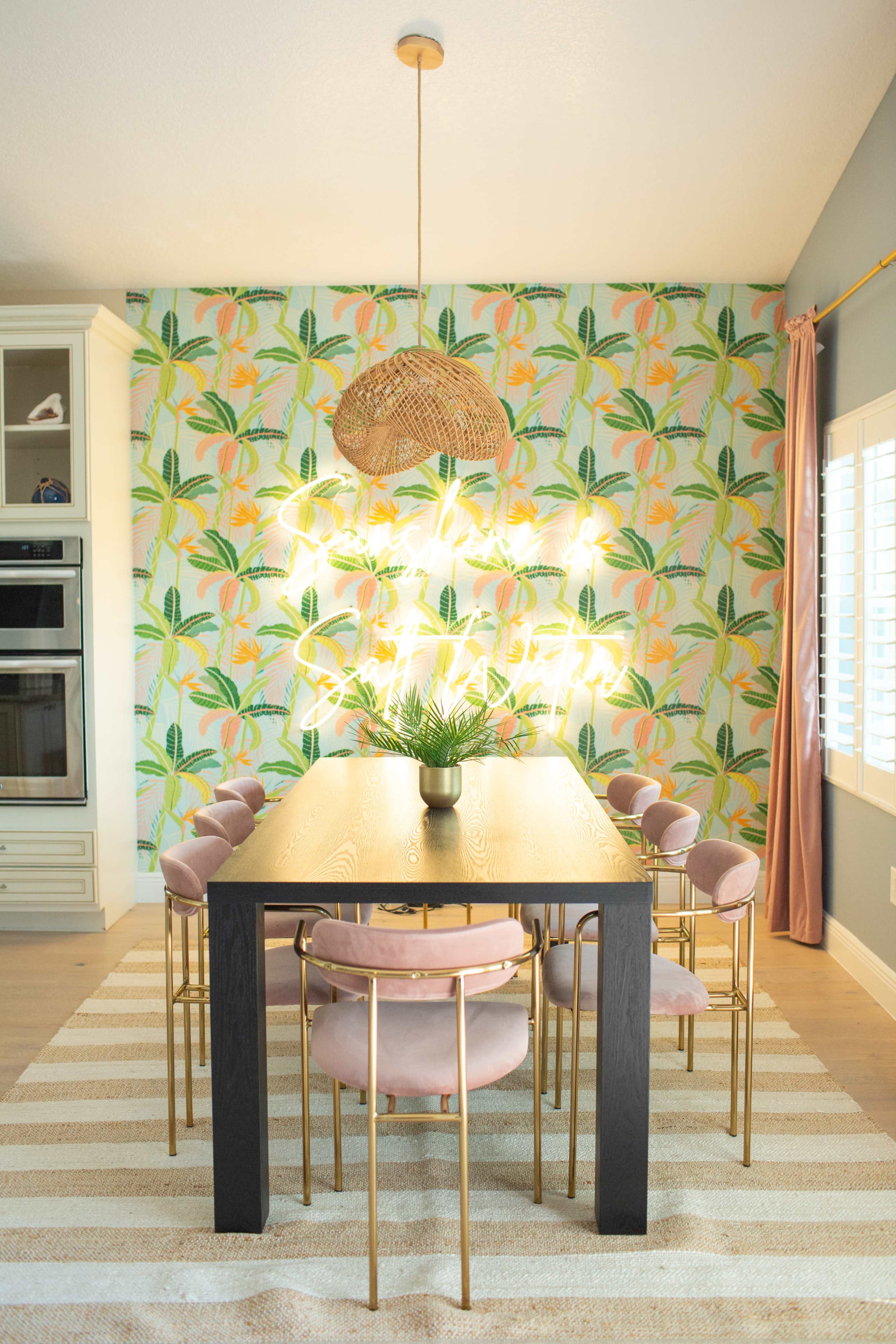 A dining area features a large black table surrounded by pink upholstered chairs, adorned with tropical-themed wallpaper and a pendant light above.