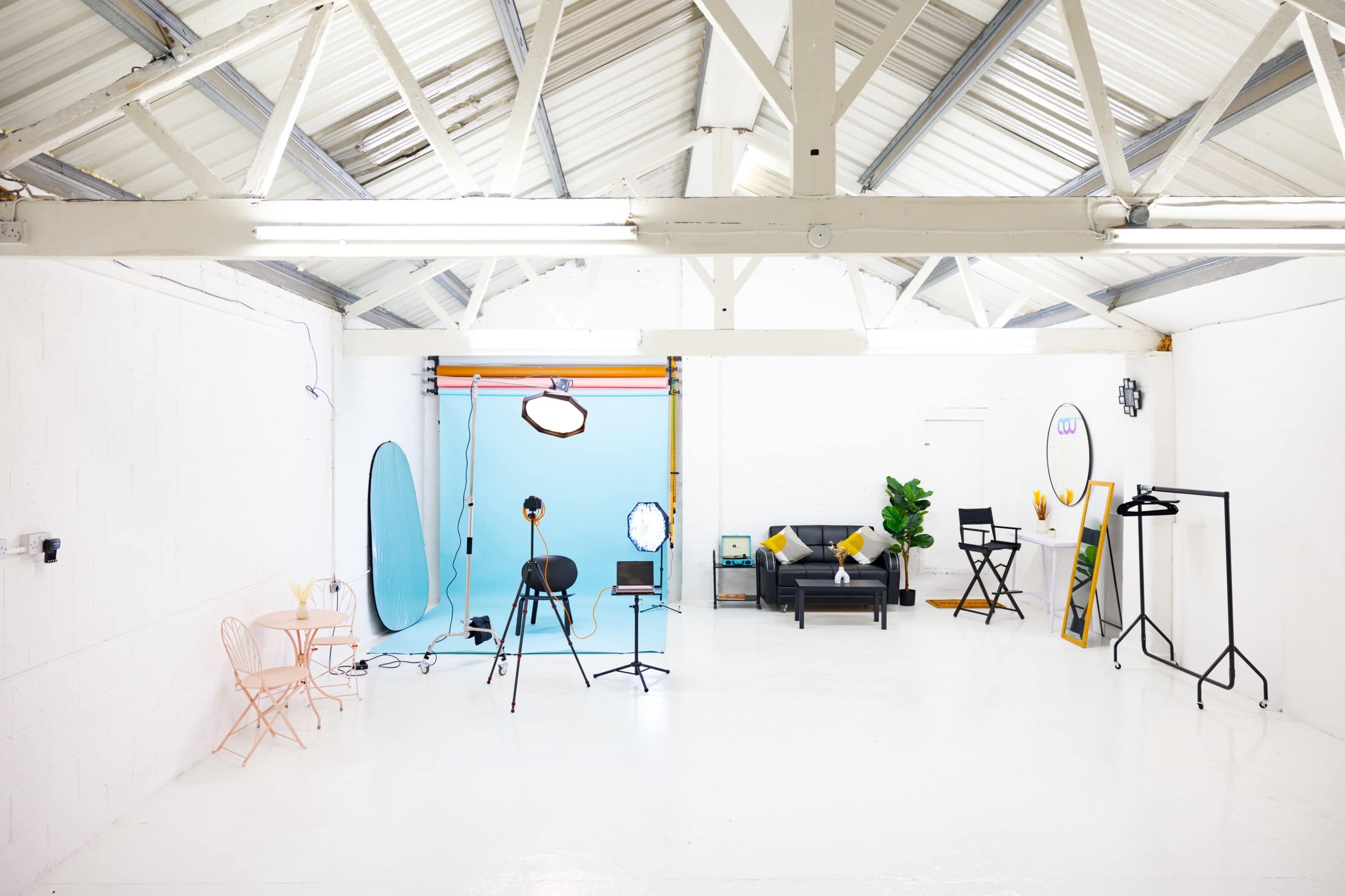 A spacious, well-lit studio features a blue backdrop, various photography equipment, and furniture including a black couch and a pink table set.
