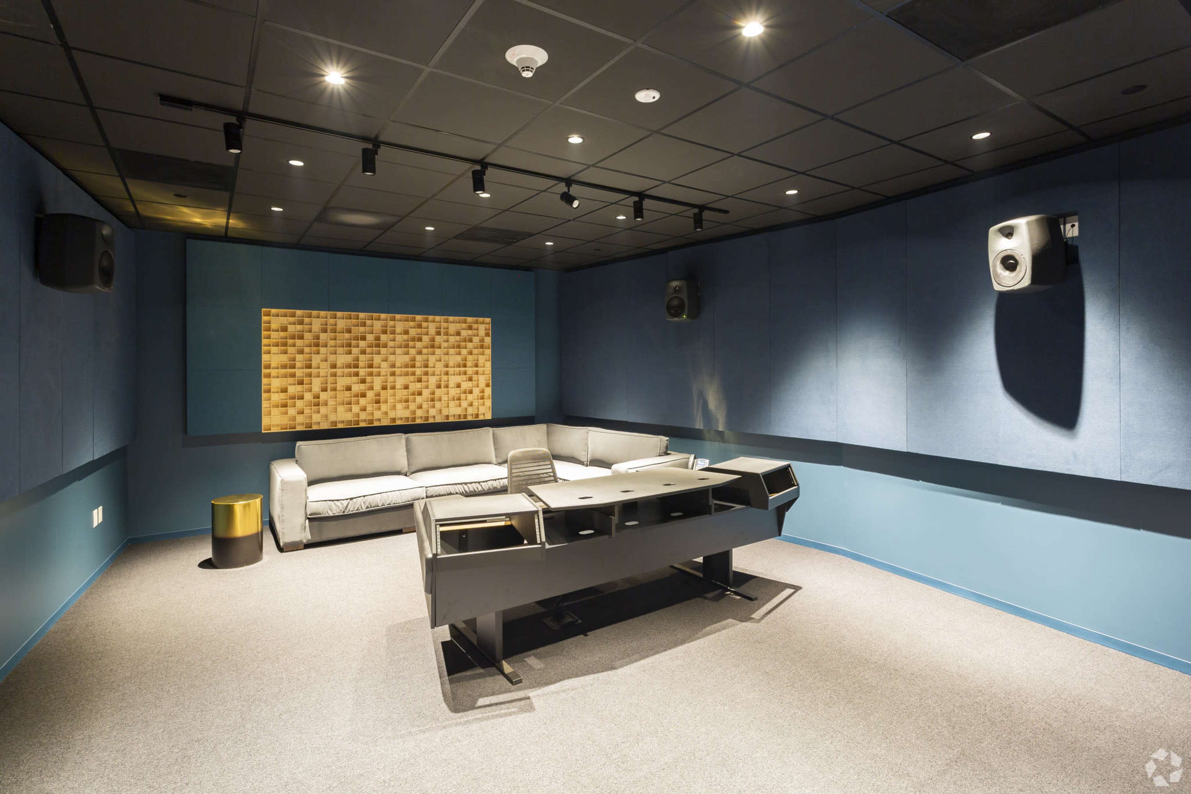 The image shows a sound studio with padded walls, a central mixing desk, a couch, and several wall-mounted speakers.