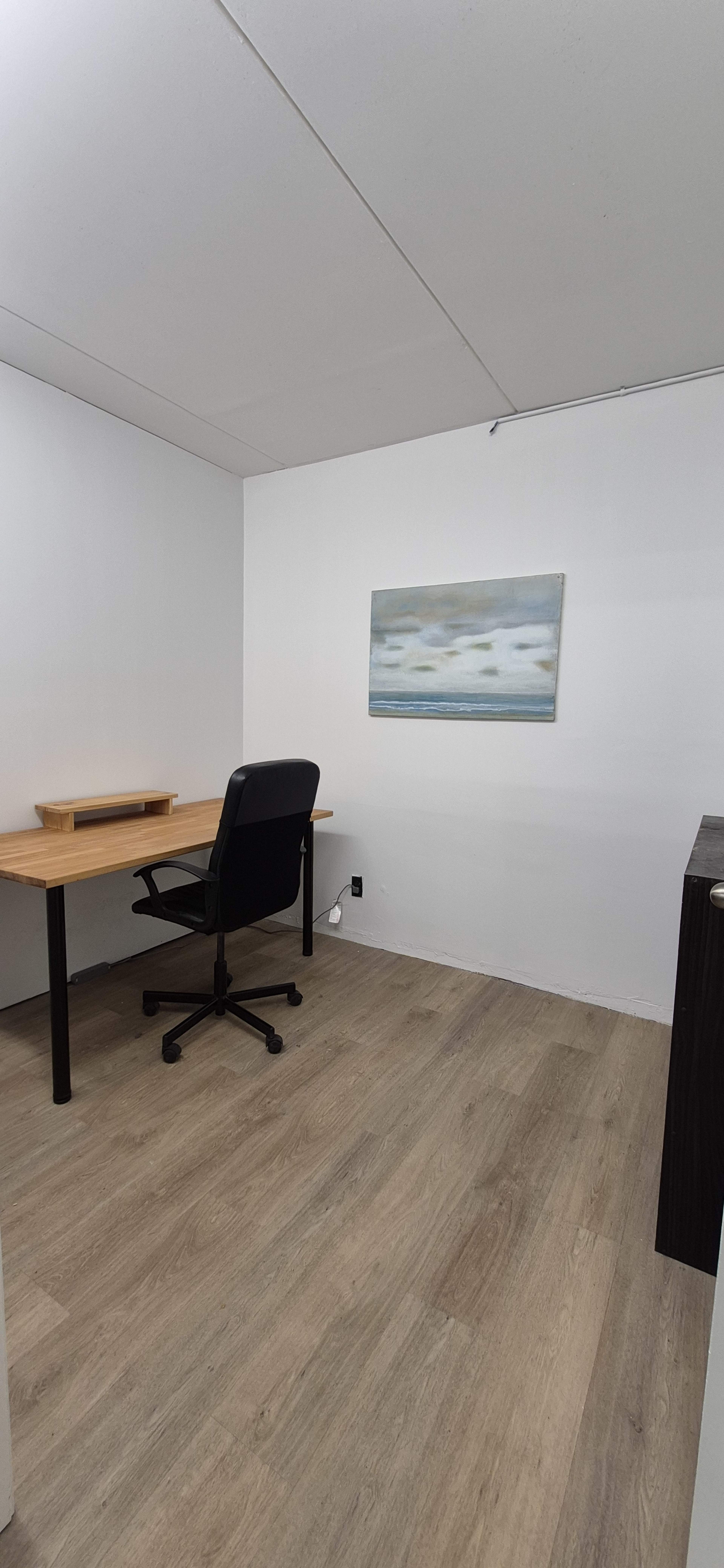 The image shows a minimalistic home office featuring a wooden desk, a black office chair, and a piece of art on the wall, all set against a light-colored background with wooden flooring.