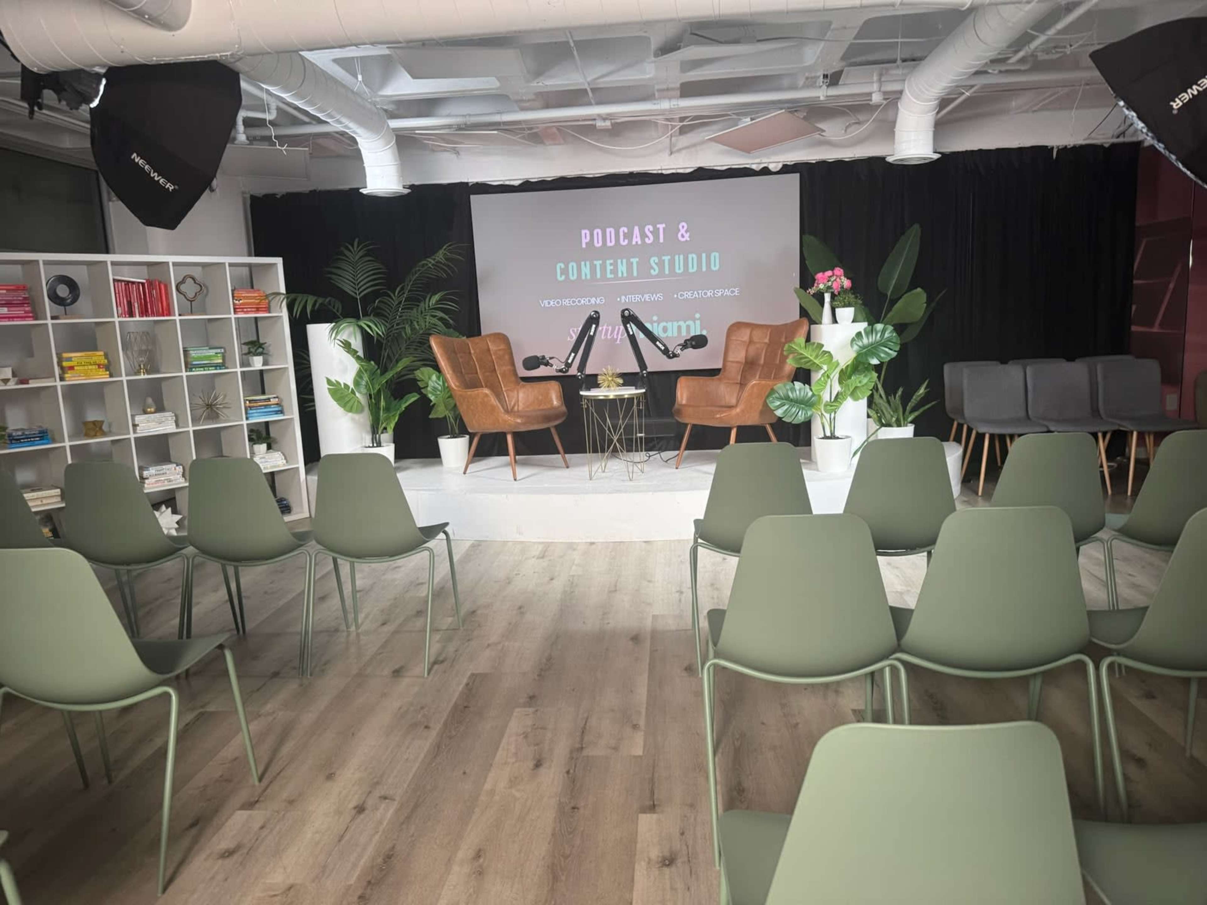 The image shows a podcast and content studio featuring two chairs, a table, greenery, and a seating area with several chairs arranged in rows.