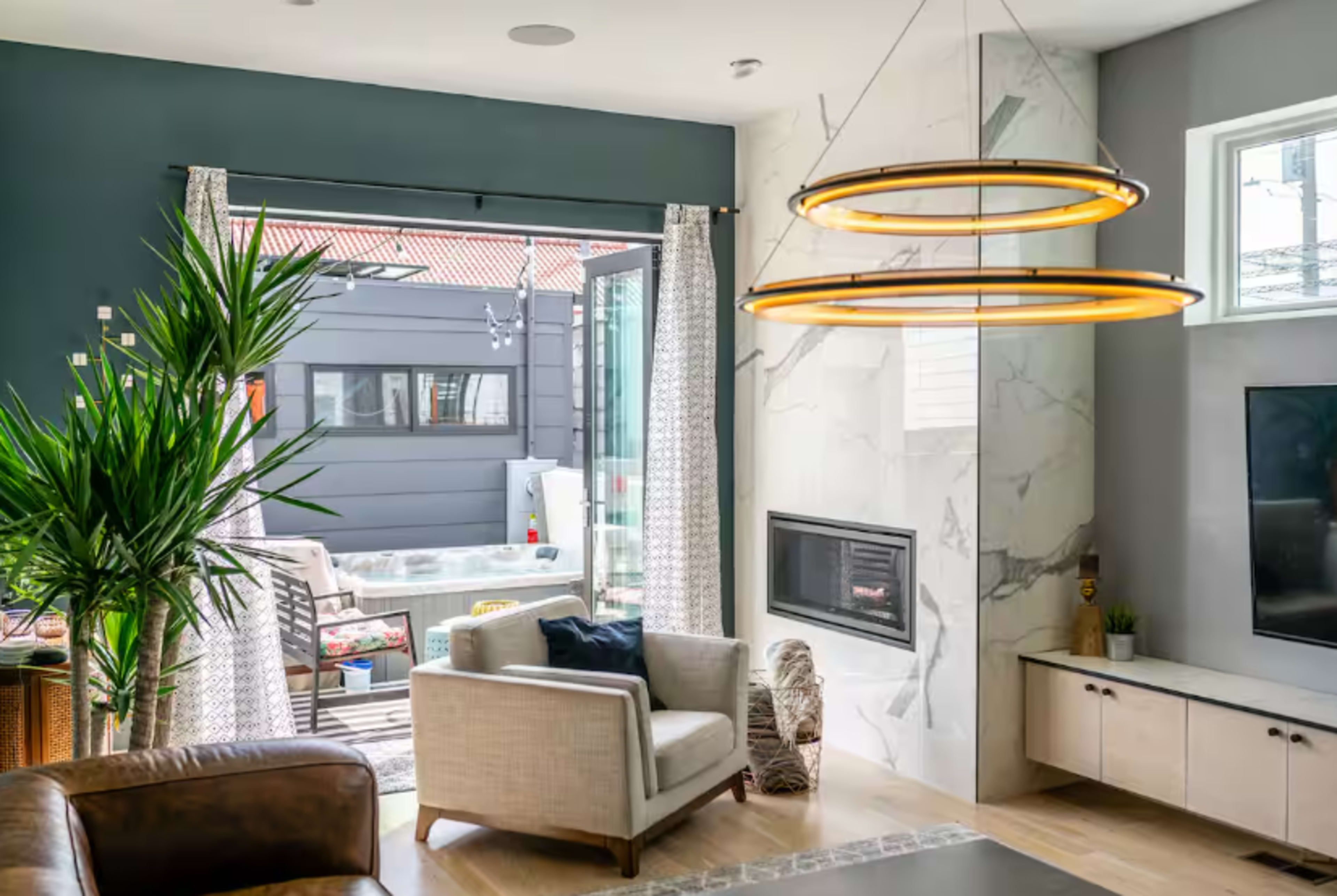 The image shows a modern living room featuring a fireplace, a large television, and a view of an outdoor patio through sliding glass doors, with stylish decor including a potted plant and pendant lighting.