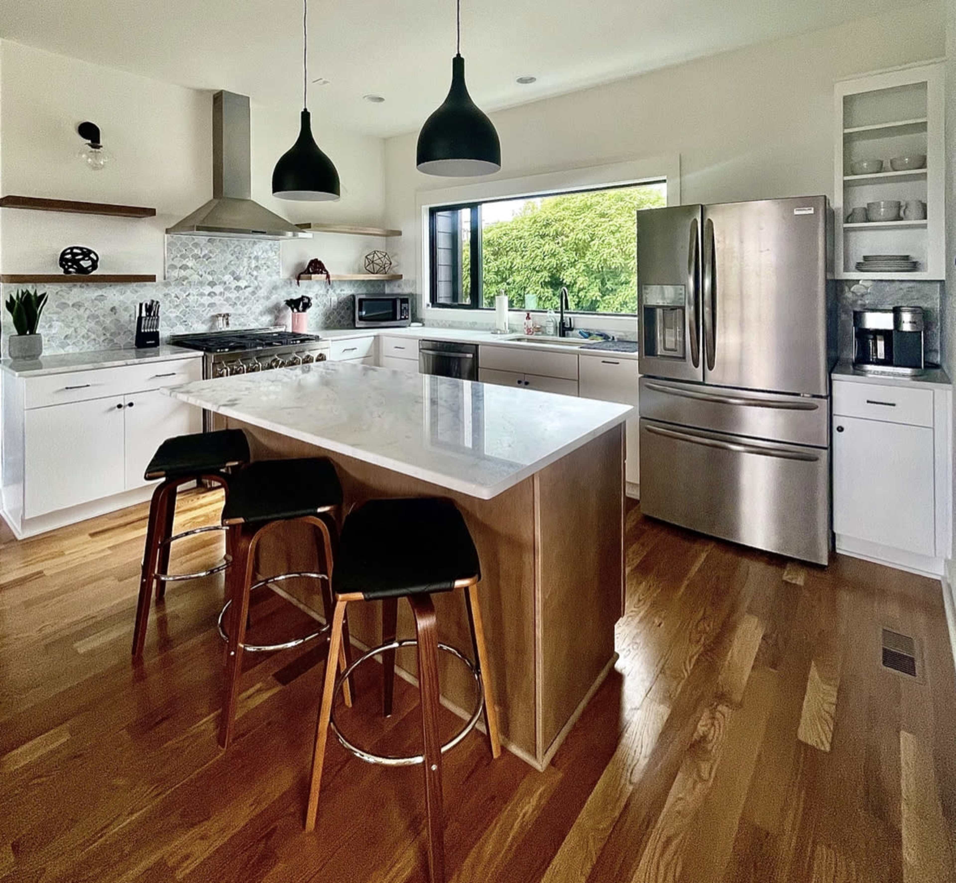 A modern kitchen features a central island with seating, stainless steel appliances, and ample natural light from a large window.