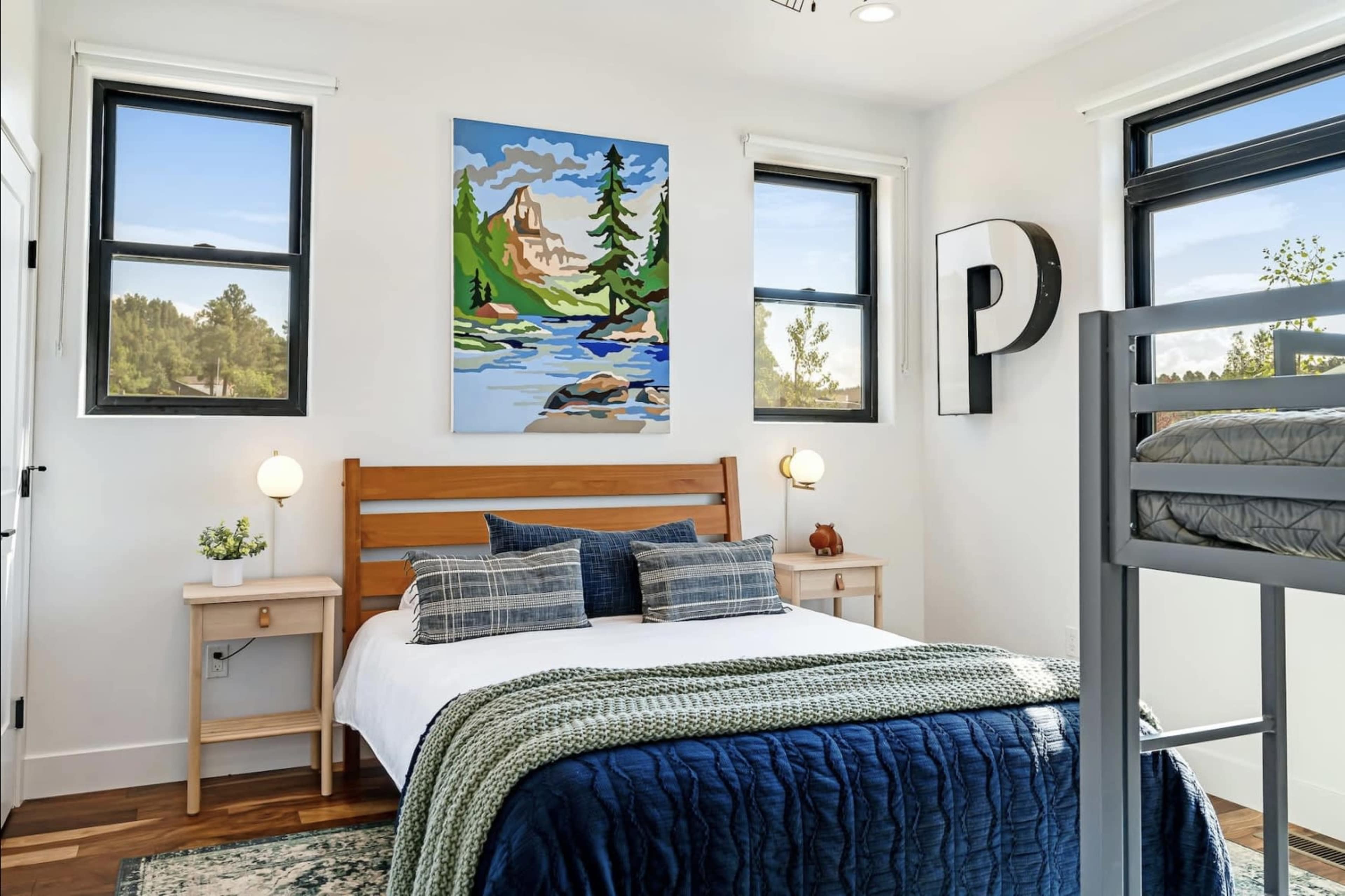 A modern bedroom features a wooden bed with a blue and white bedding set, flanked by two nightstands and adorned with a large landscape painting above the bed.