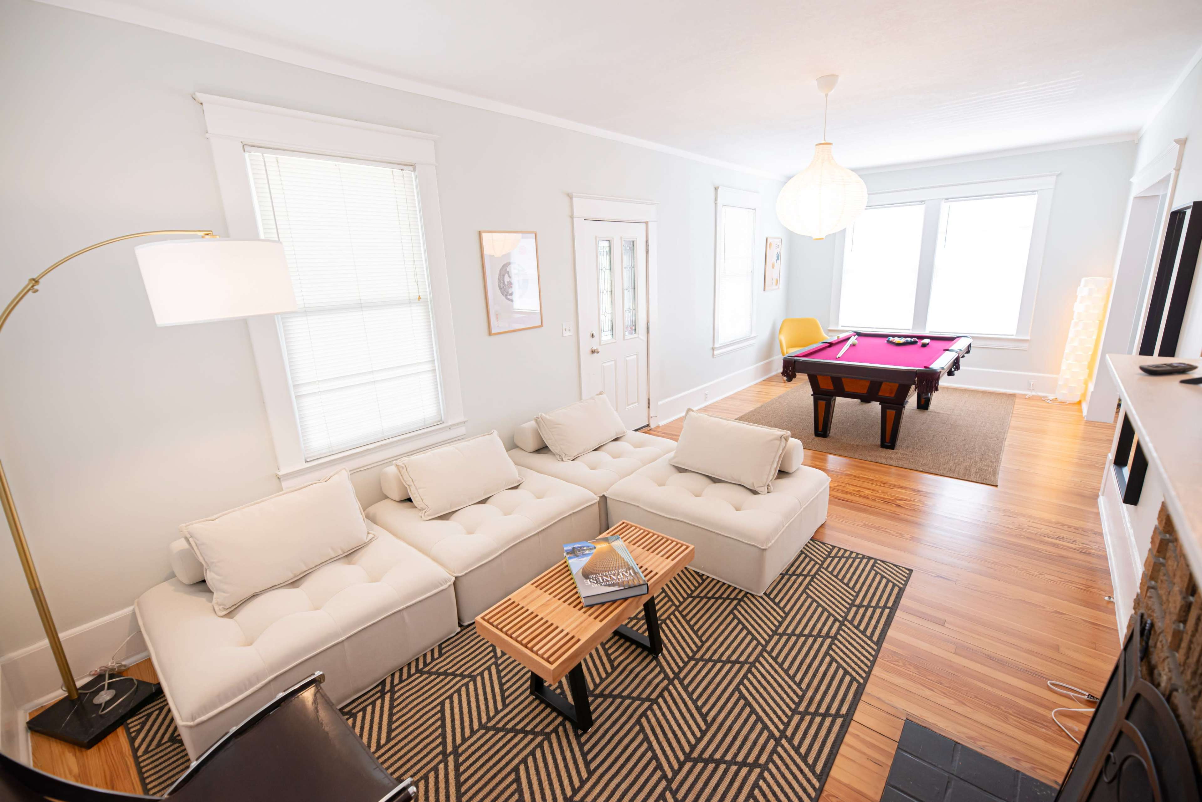 A well-lit living room featuring a light-colored sectional sofa, a pool table, and large windows with sheer curtains.